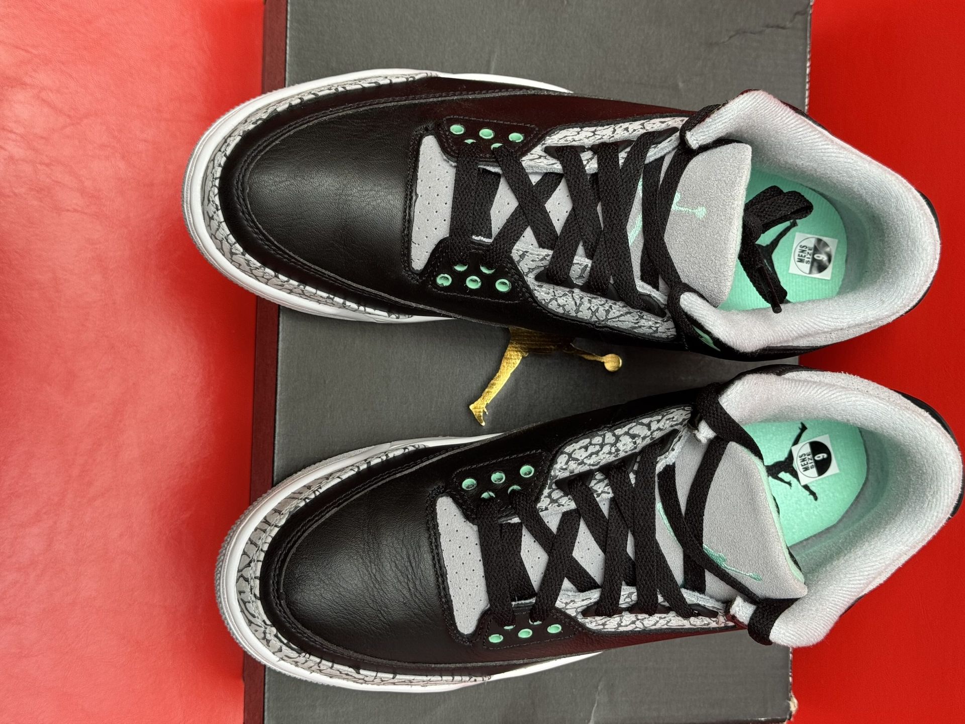 Black and gray Air Jordan 3 sneakers on a black box, one shoe upside down showing its sole; red background.