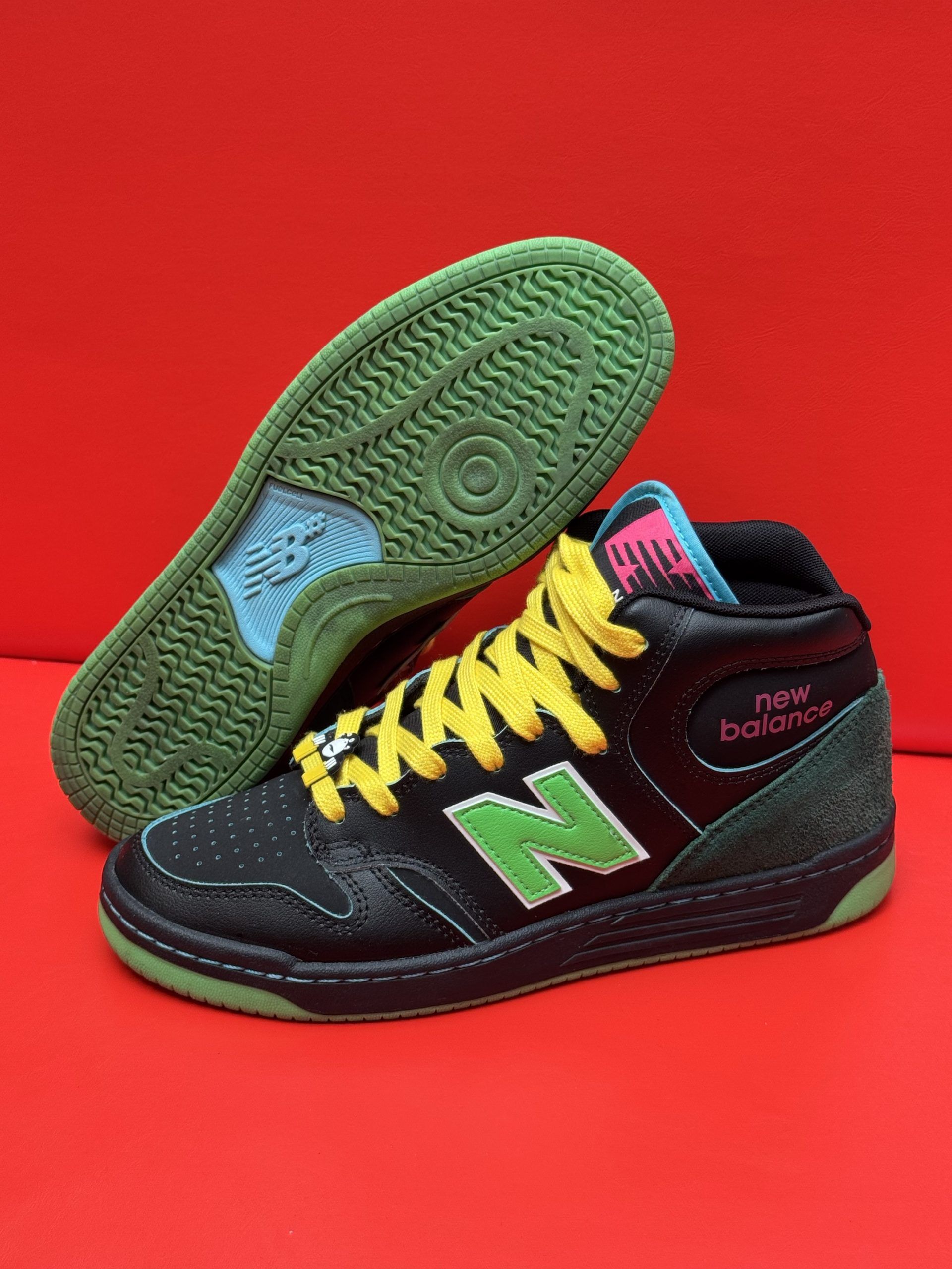 Black and green New Balance high-top sneaker with yellow laces, sole visible on red background.