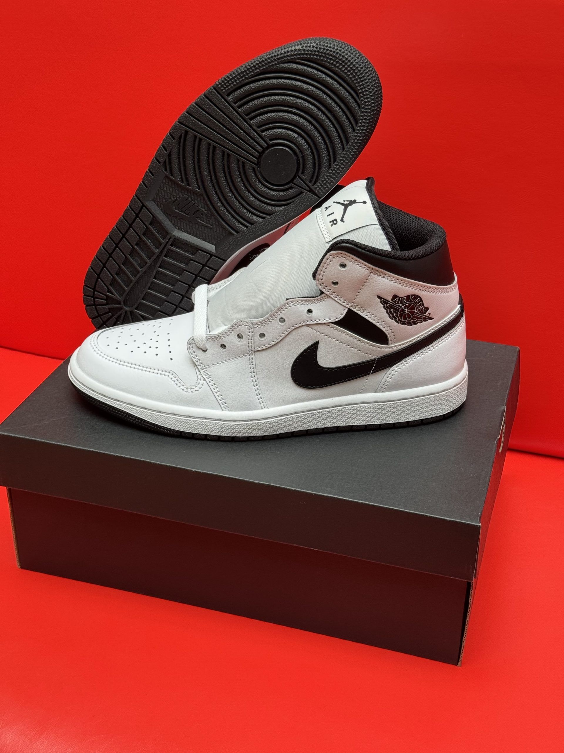 White and black Air Jordan 1 sneaker on a black box, sole visible. Red background.