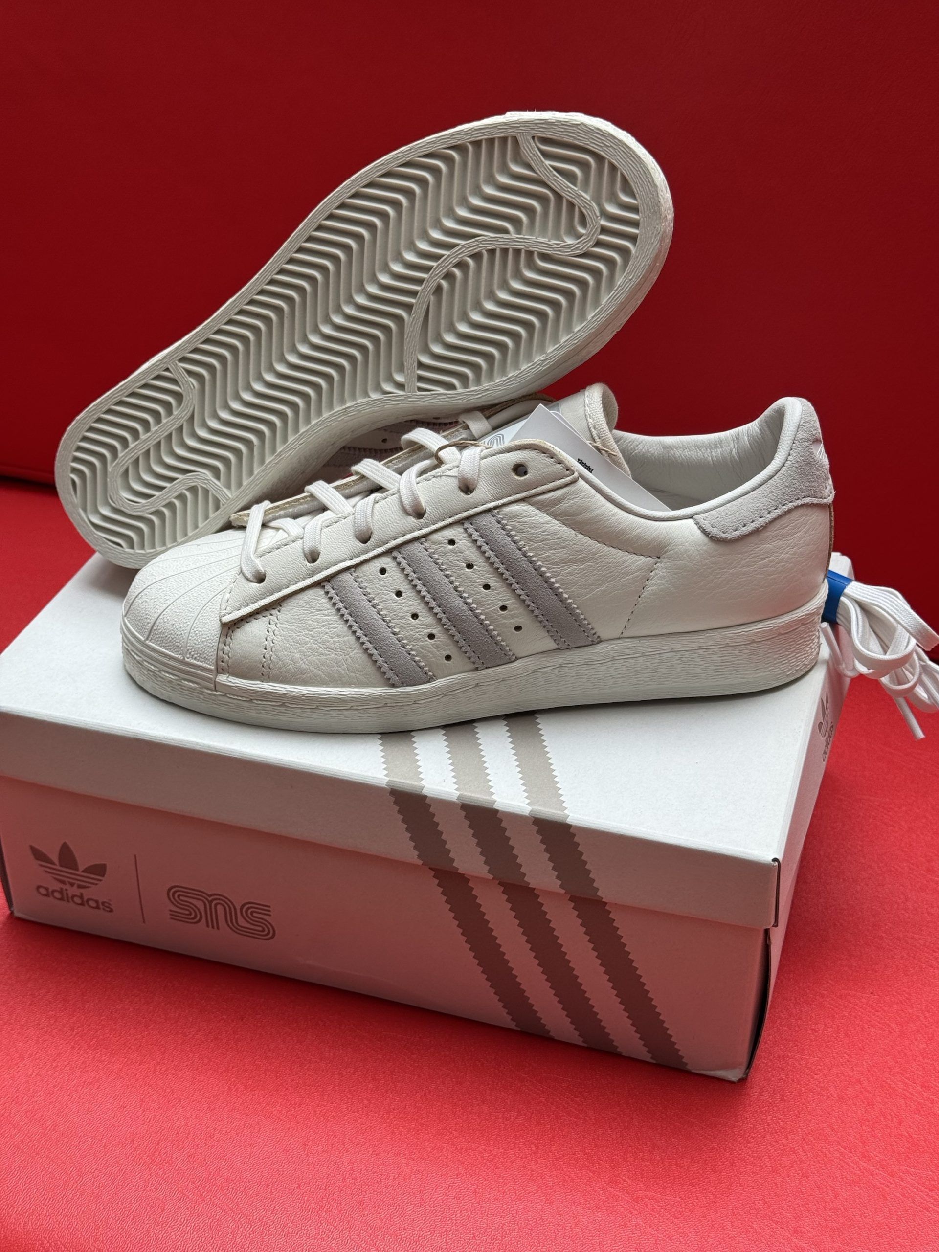 White Adidas Superstar sneakers with light purple stripes, on a box, sole visible.