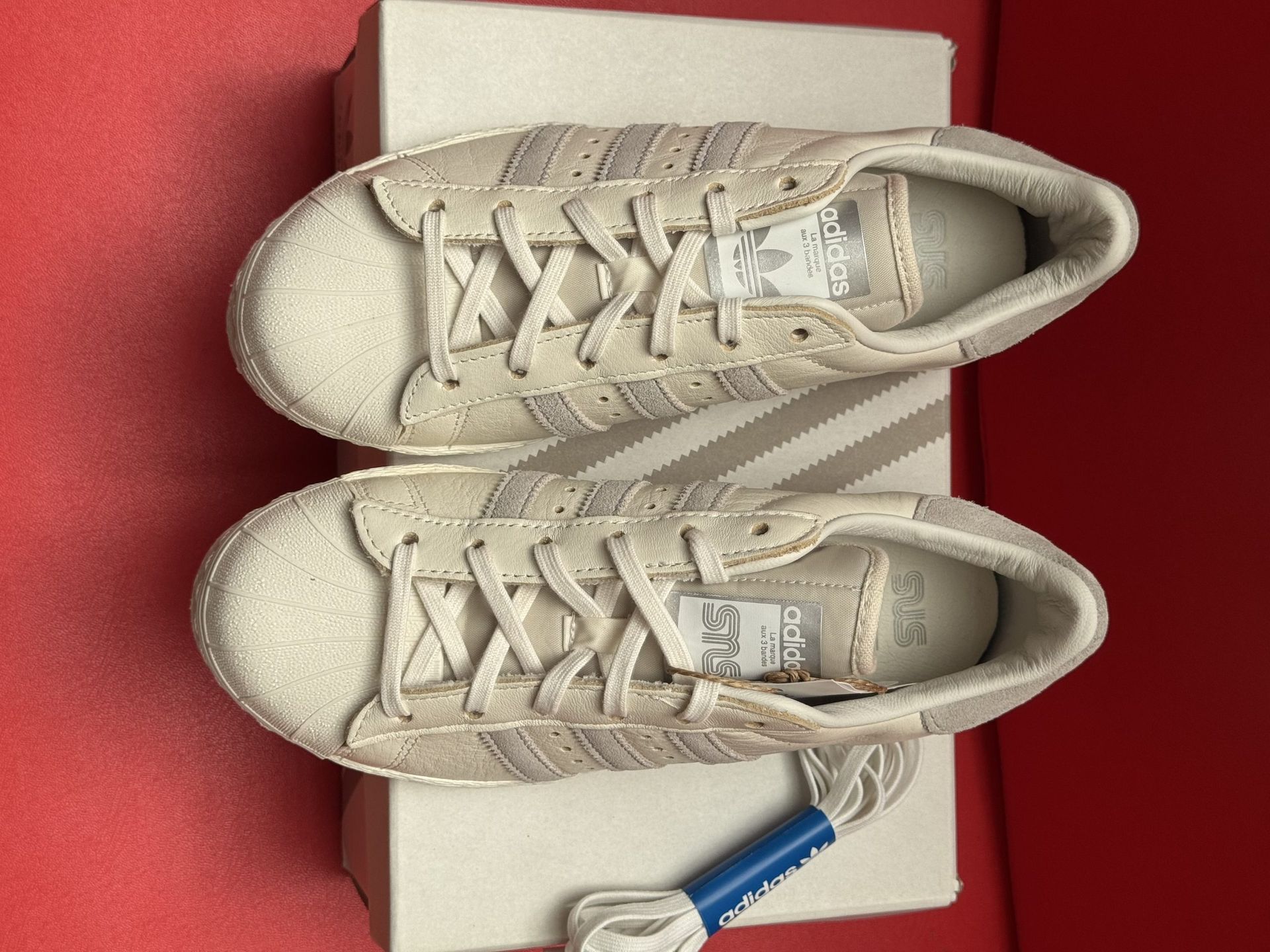 White Adidas Superstar sneakers with light purple stripes, on a box, sole visible.