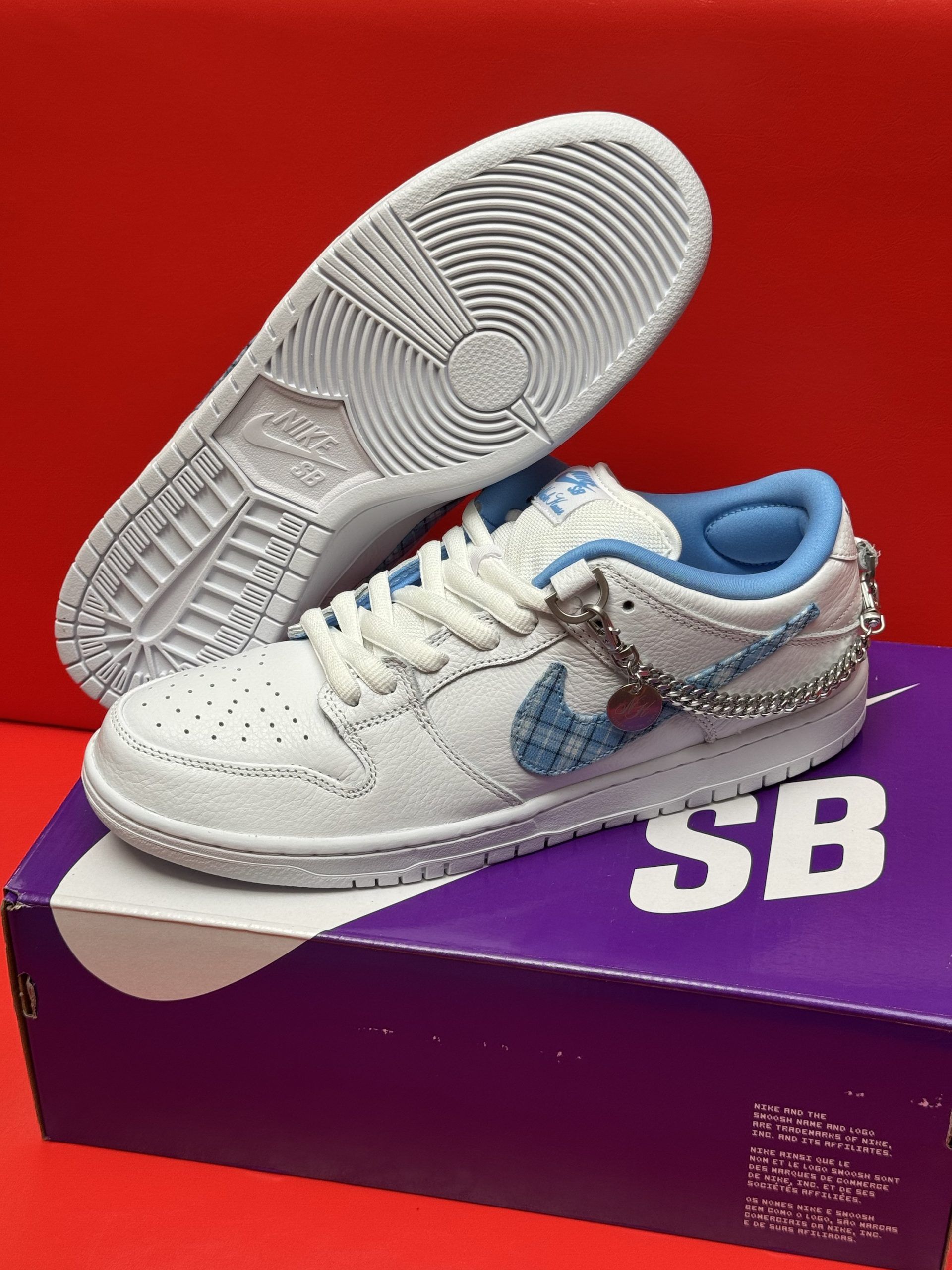 White Nike SB Dunk Low sneakers with blue accents, on a purple box against a red background.