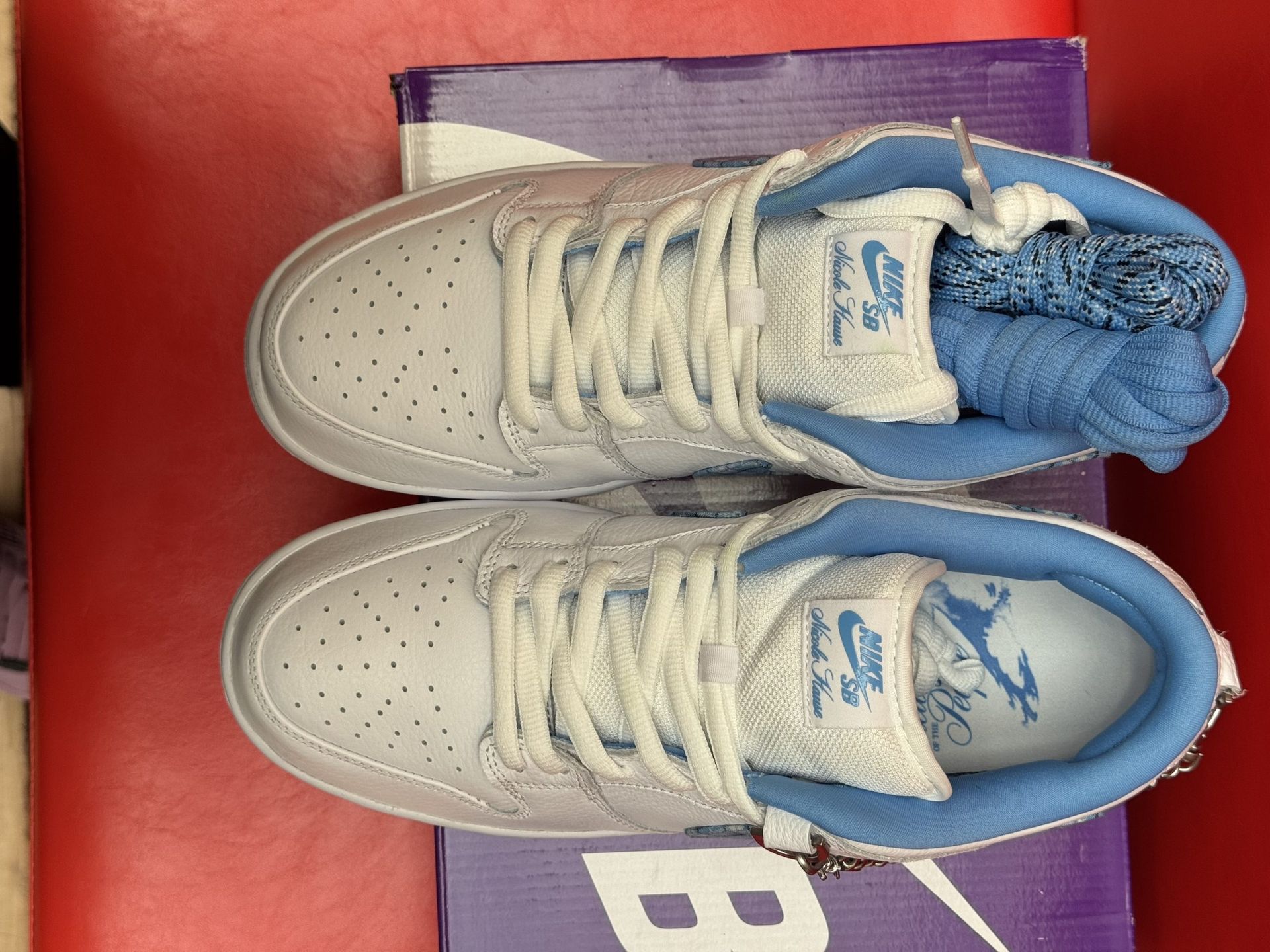 White Nike SB Dunk Low sneakers with blue accents, on a purple box against a red background.