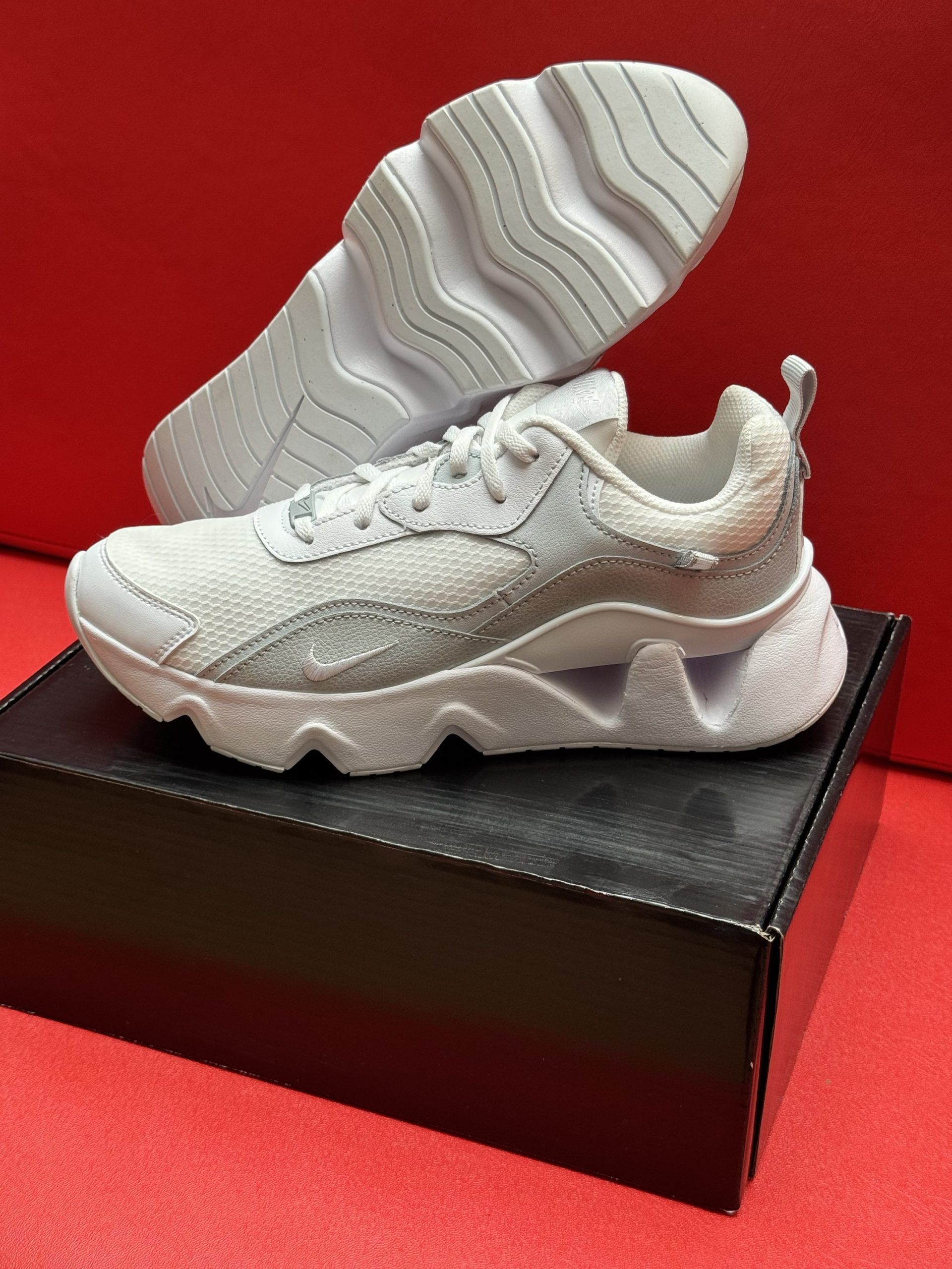 White sneakers on a black box, one showing its wavy sole. Red background.