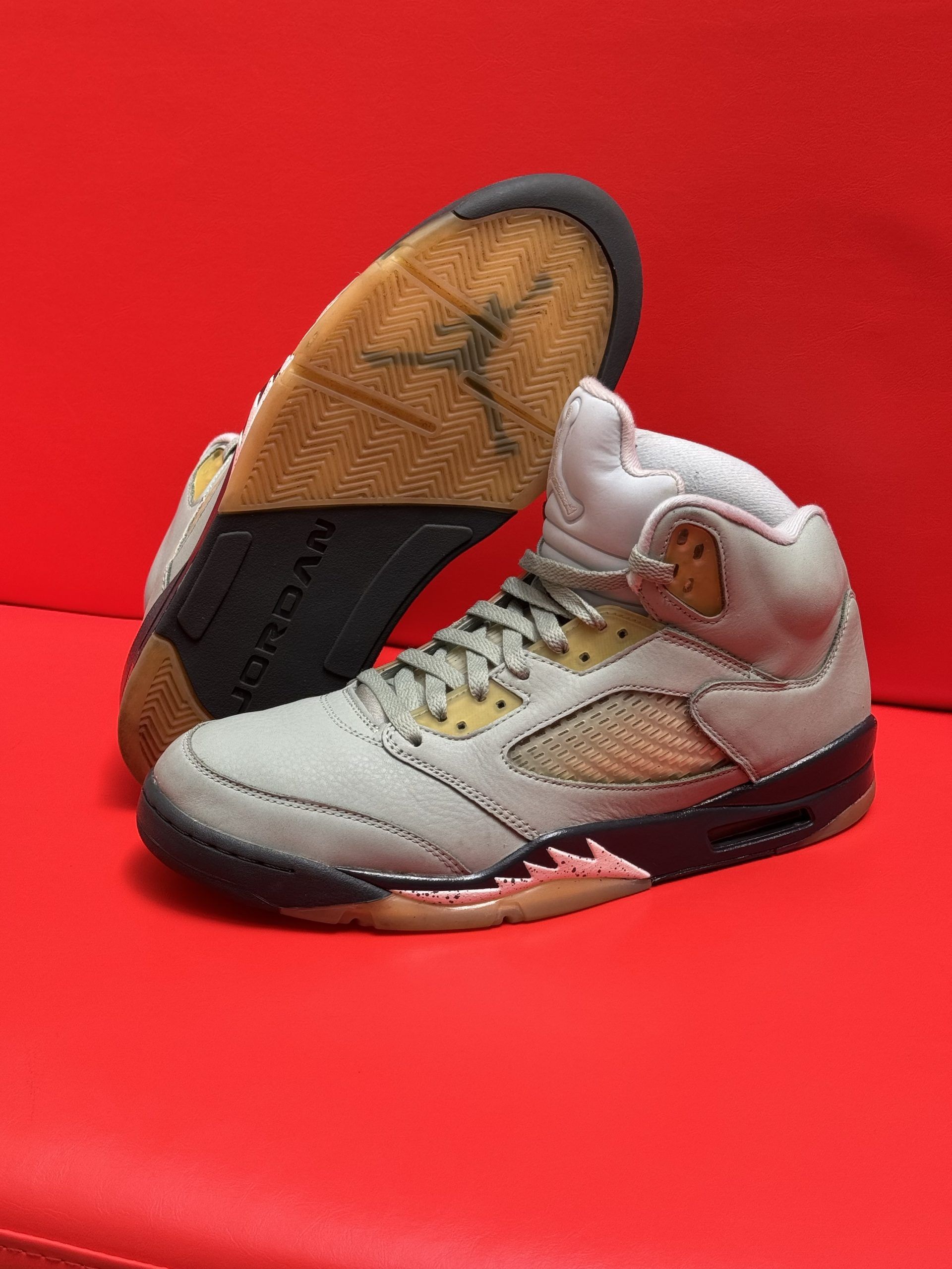 Gray, tan, and black Air Jordan 5 sneakers, one upside down showing the sole, on a red surface.