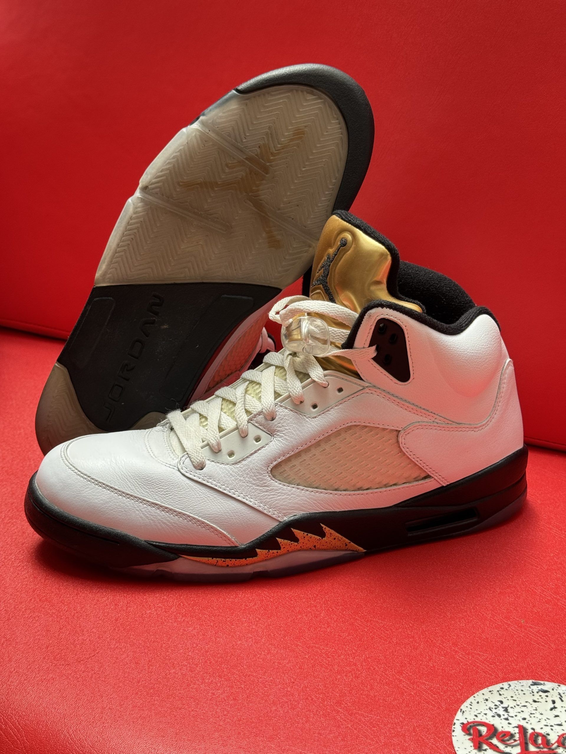 White, black, and gold Air Jordan 5 sneaker with a worn outsole, set against a red background.