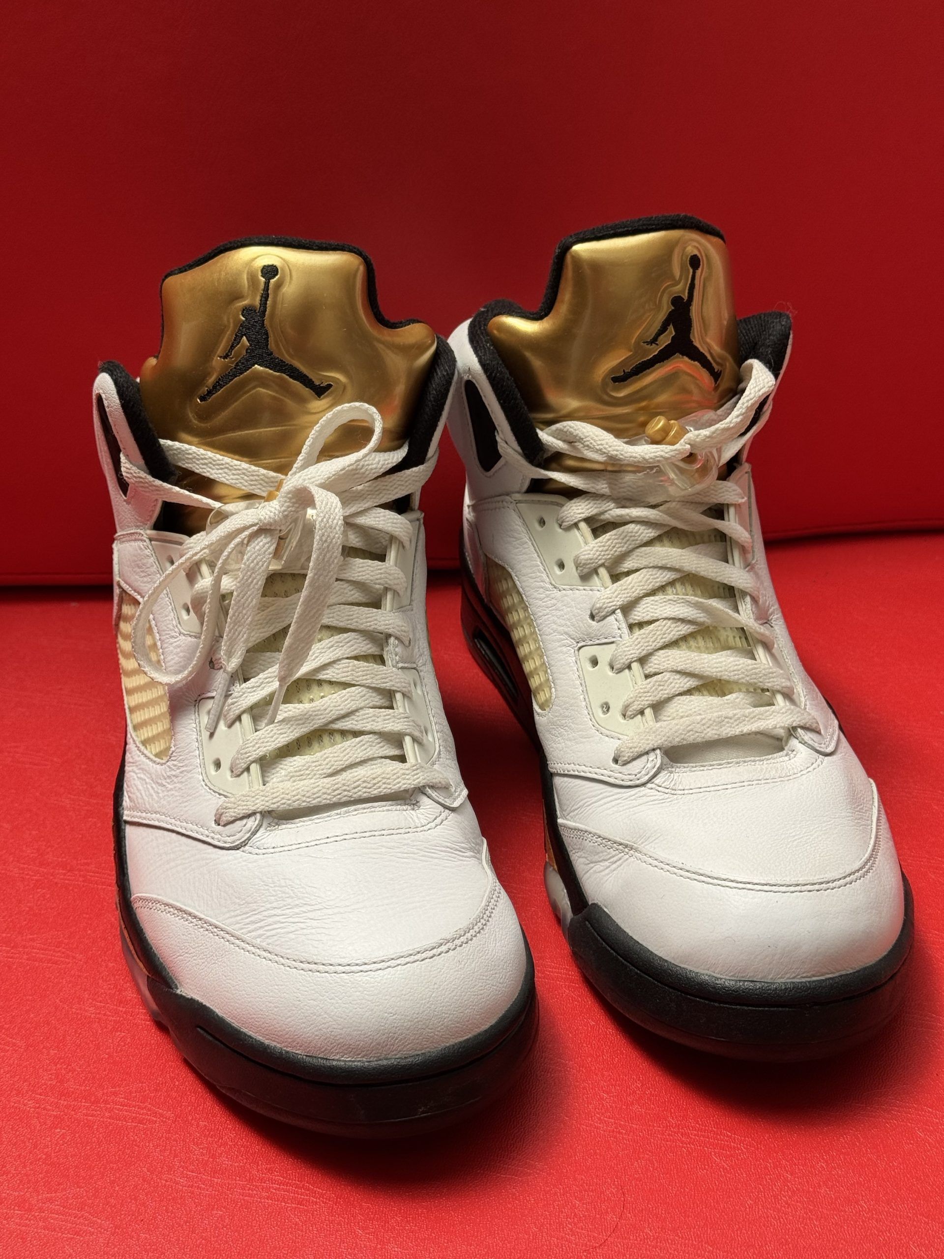 White, black, and gold Air Jordan 5 sneaker with a worn outsole, set against a red background.