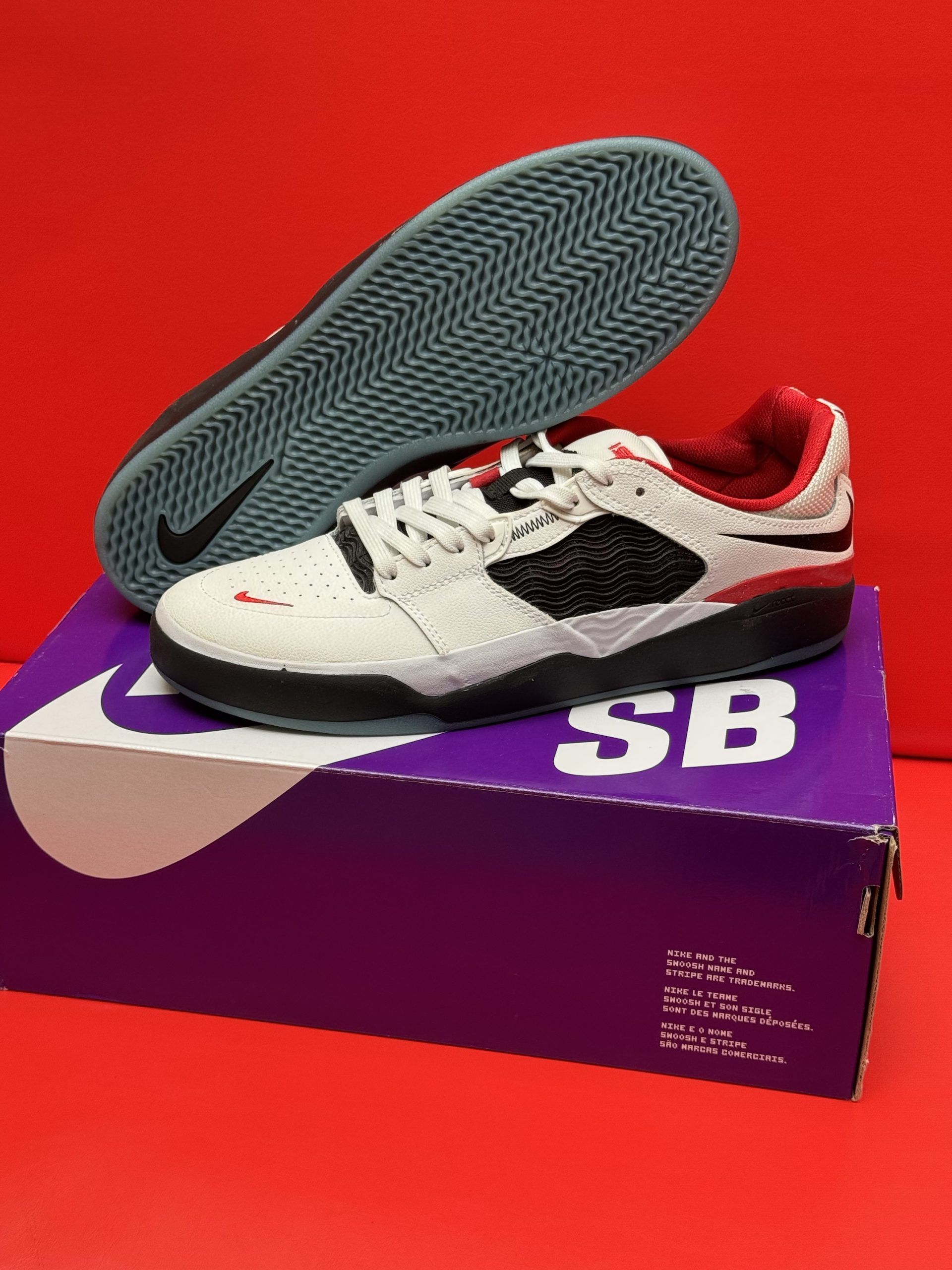 White and red Nike SB sneakers on a purple box against a red backdrop, sole visible.
