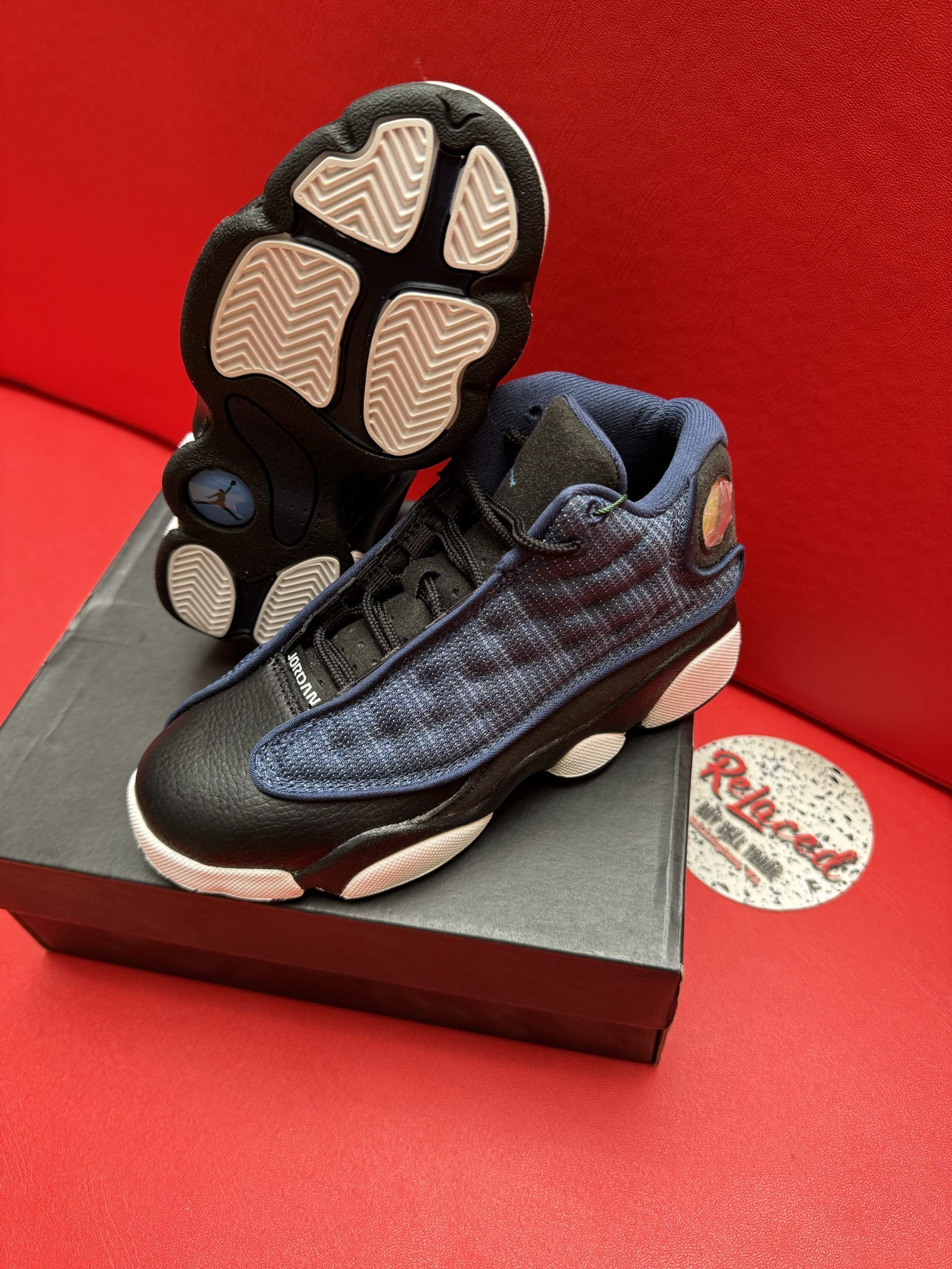 Pair of toddler Air Jordan 13 sneakers, black leather and blue-patterned uppers, white soles, resting on a black box, against a red background.