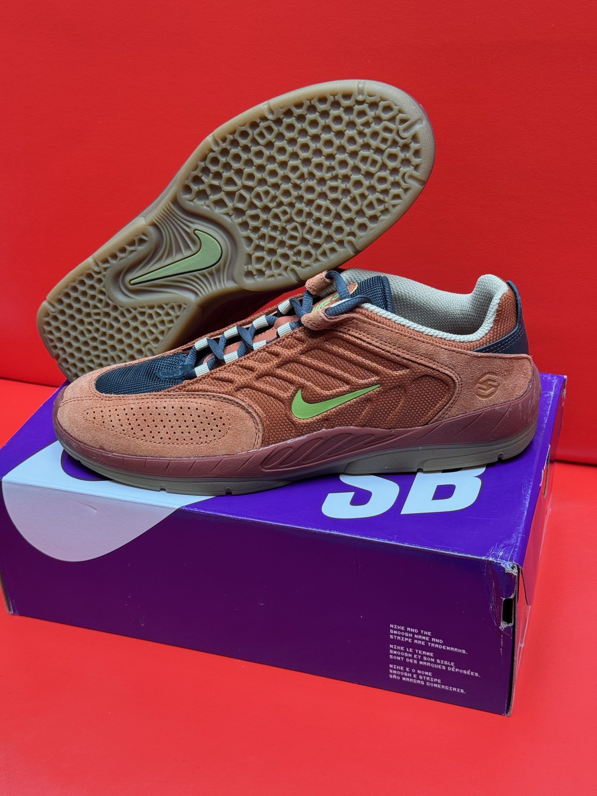 Nike SB Dunk Low Pro sneakers in orange and brown, on a purple SB box, with a red background.