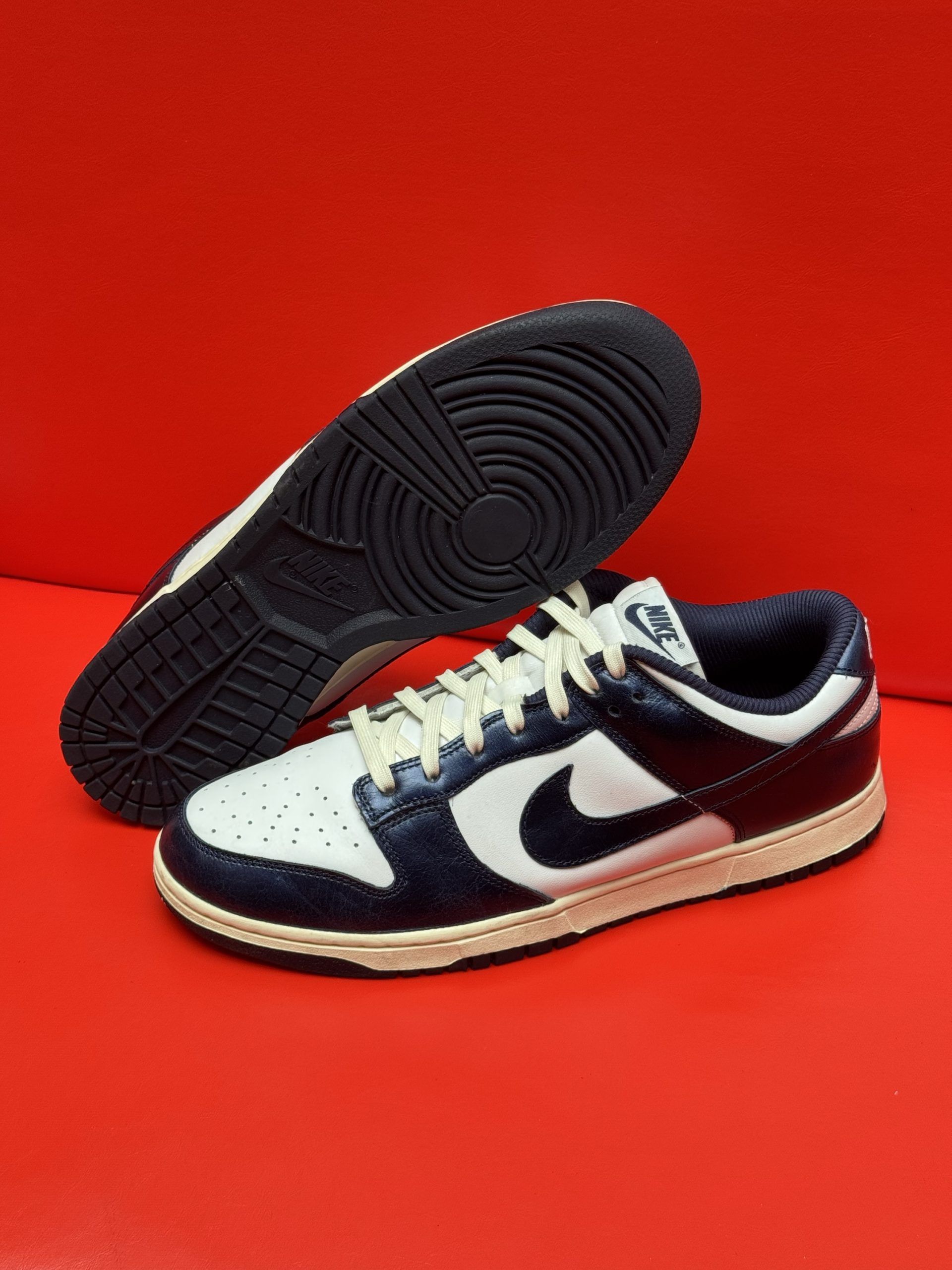 White and navy Nike Dunk Low sneakers, one showing sole, set against a red background.