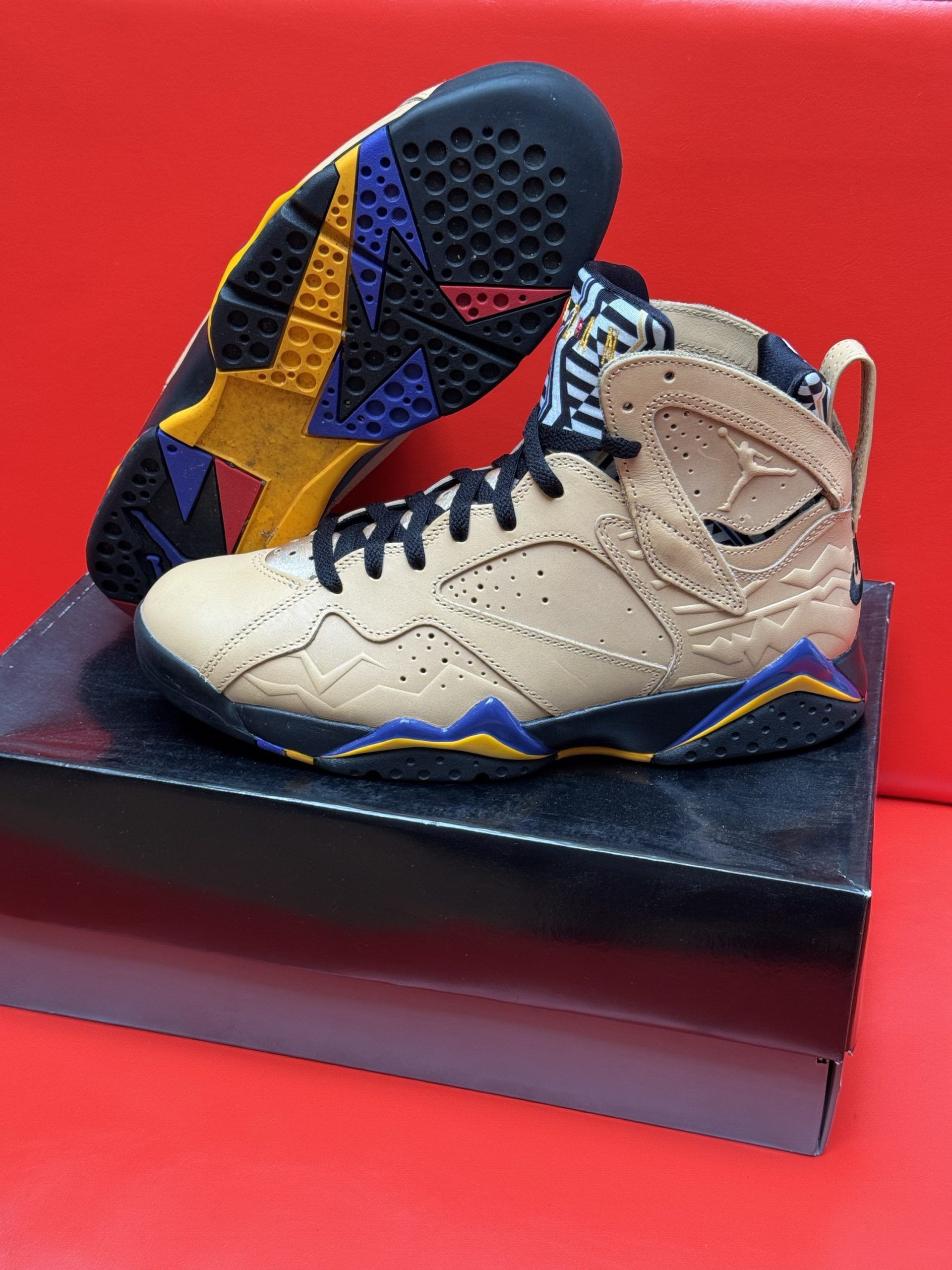 Tan and black Air Jordan 7 sneakers on a black box, sole visible, against a red background.
