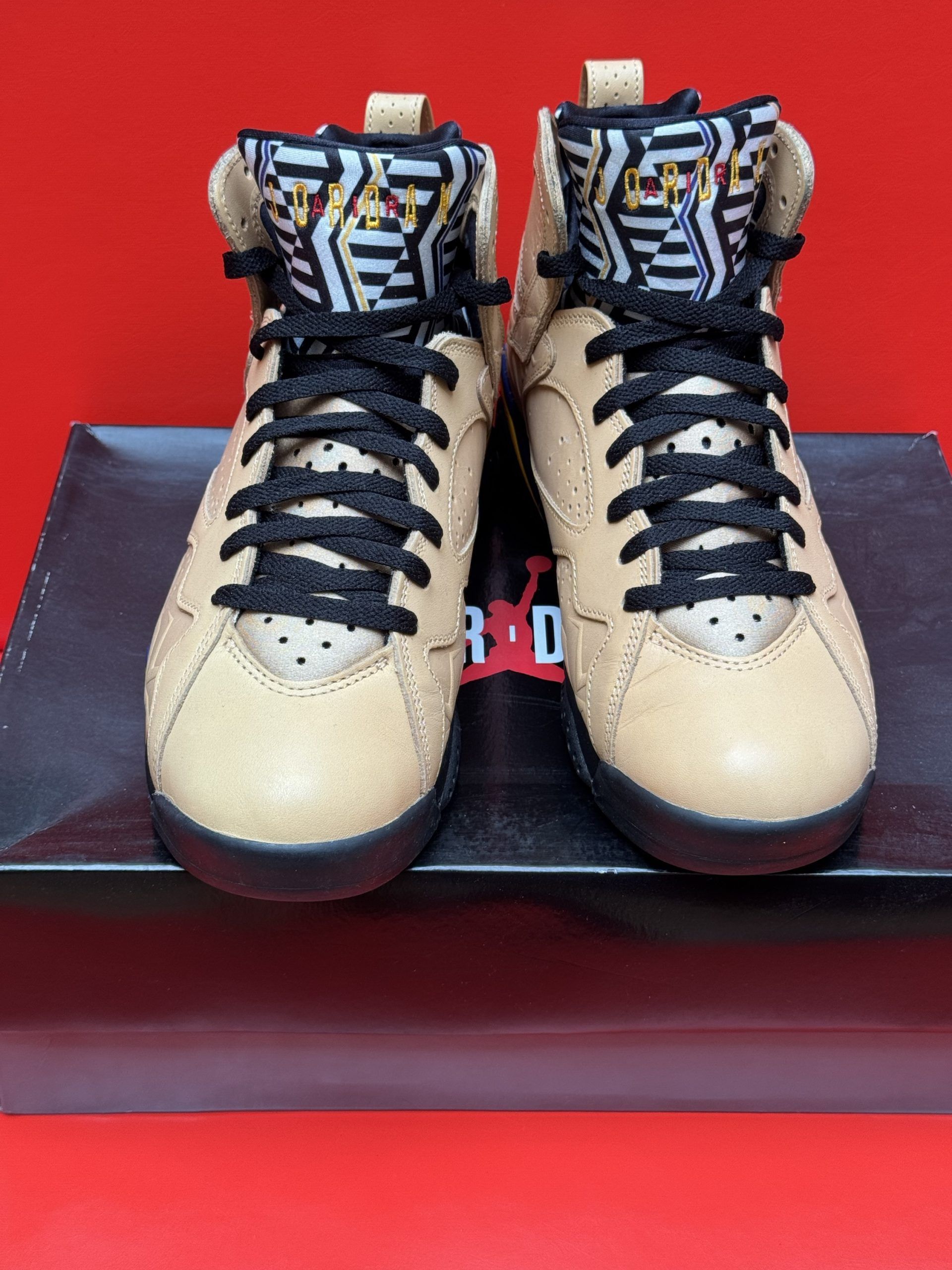 Tan and black Air Jordan 7 sneakers on a black box, sole visible, against a red background.