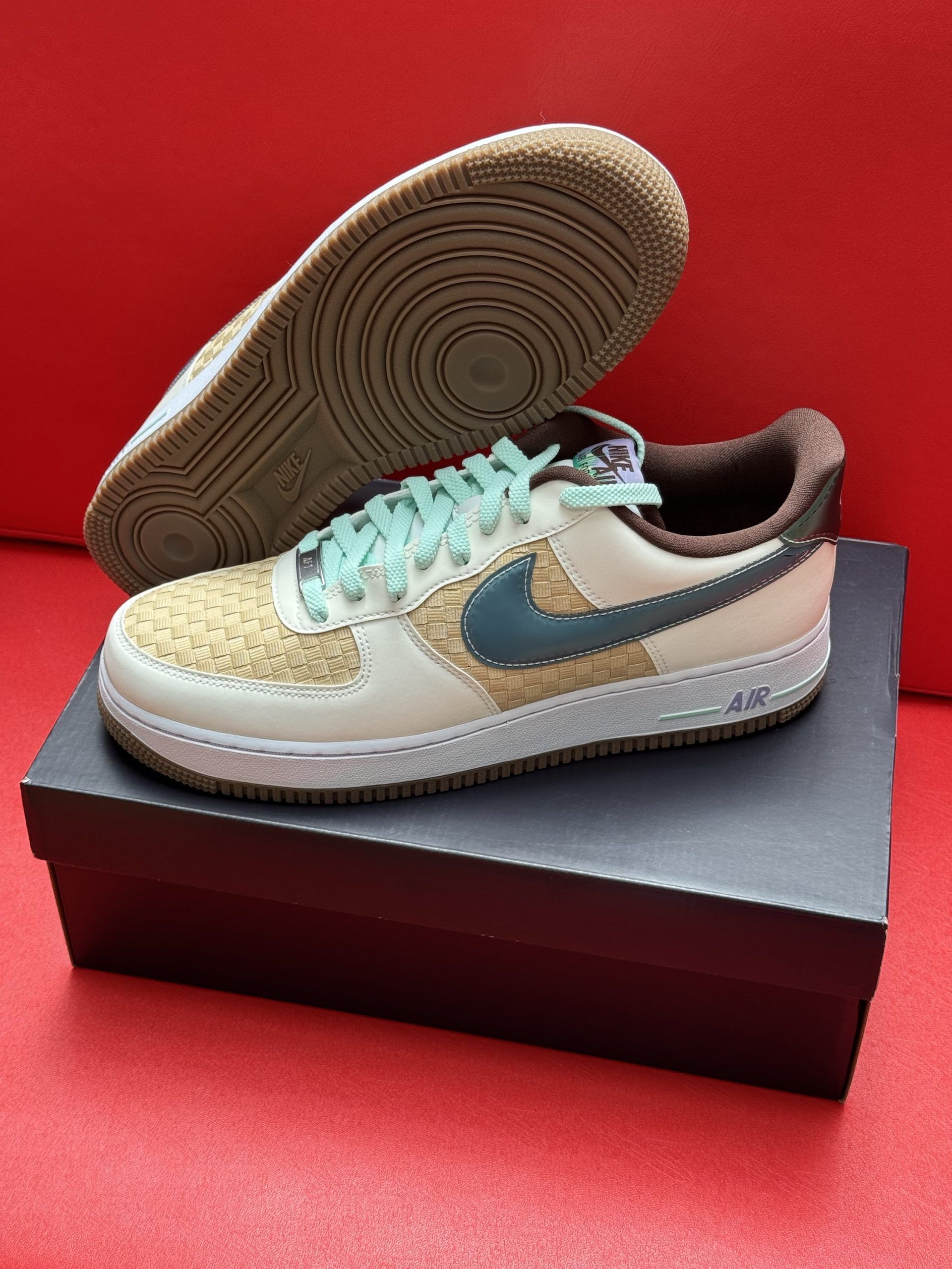 Nike Air Force 1 sneaker on a black box, displayed on a red surface. The shoe is beige, blue and brown.