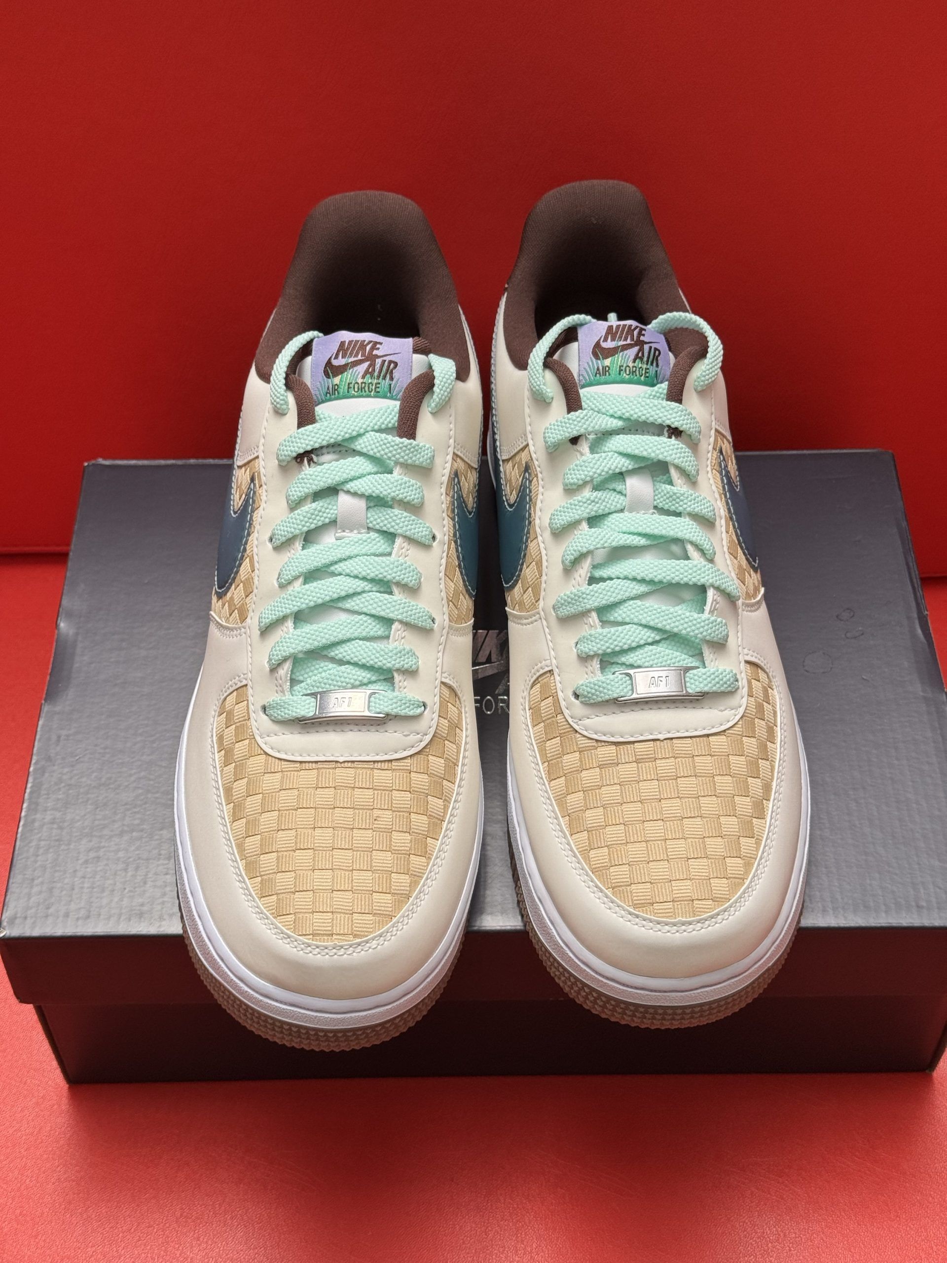 Nike Air Force 1 sneaker on a black box, displayed on a red surface. The shoe is beige, blue and brown.