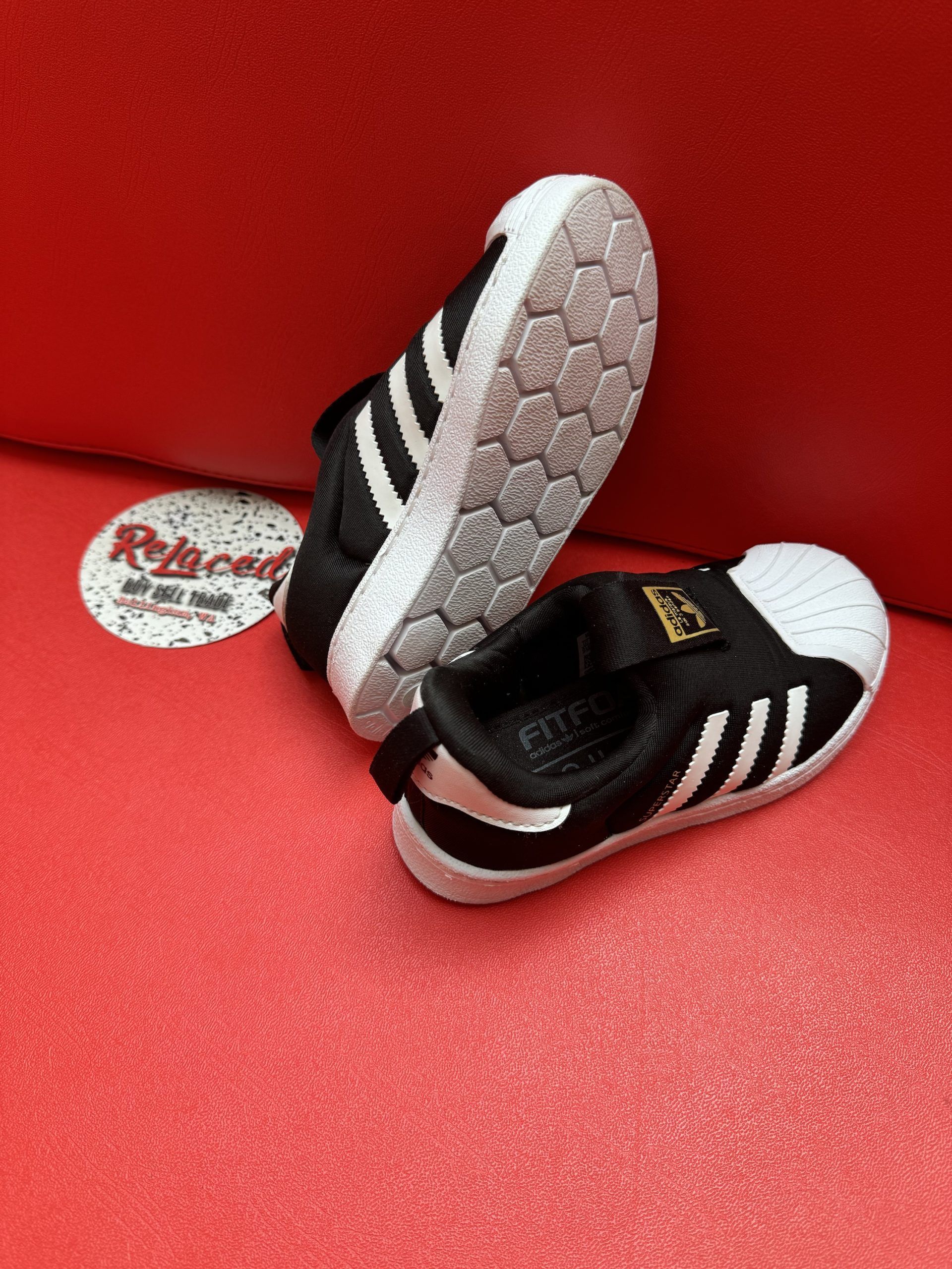 Black and white Adidas Superstar infant shoes on a red surface.