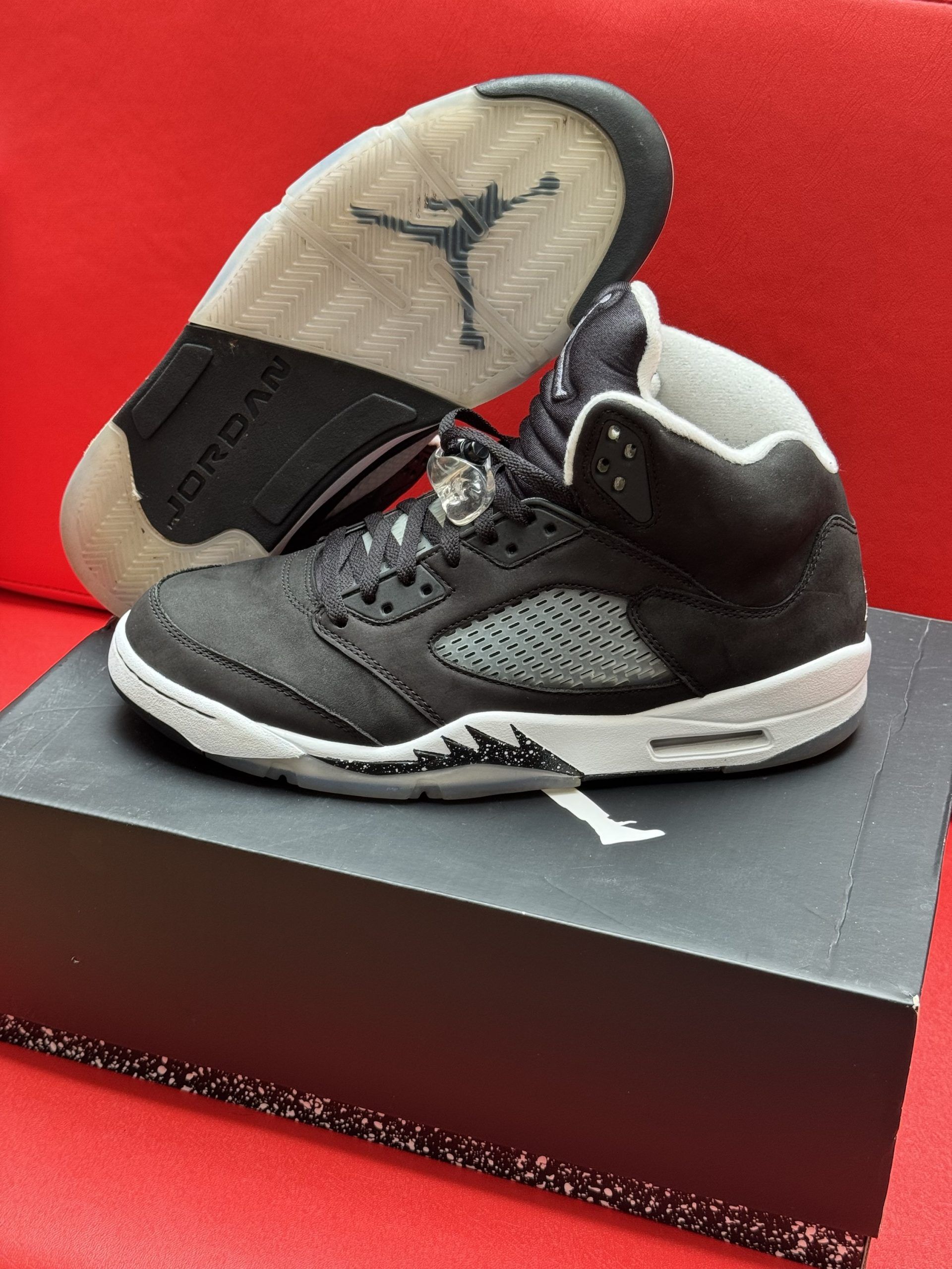 Black and white Air Jordan 5 sneakers with a clear sole, sitting on a black shoebox, against a red background.