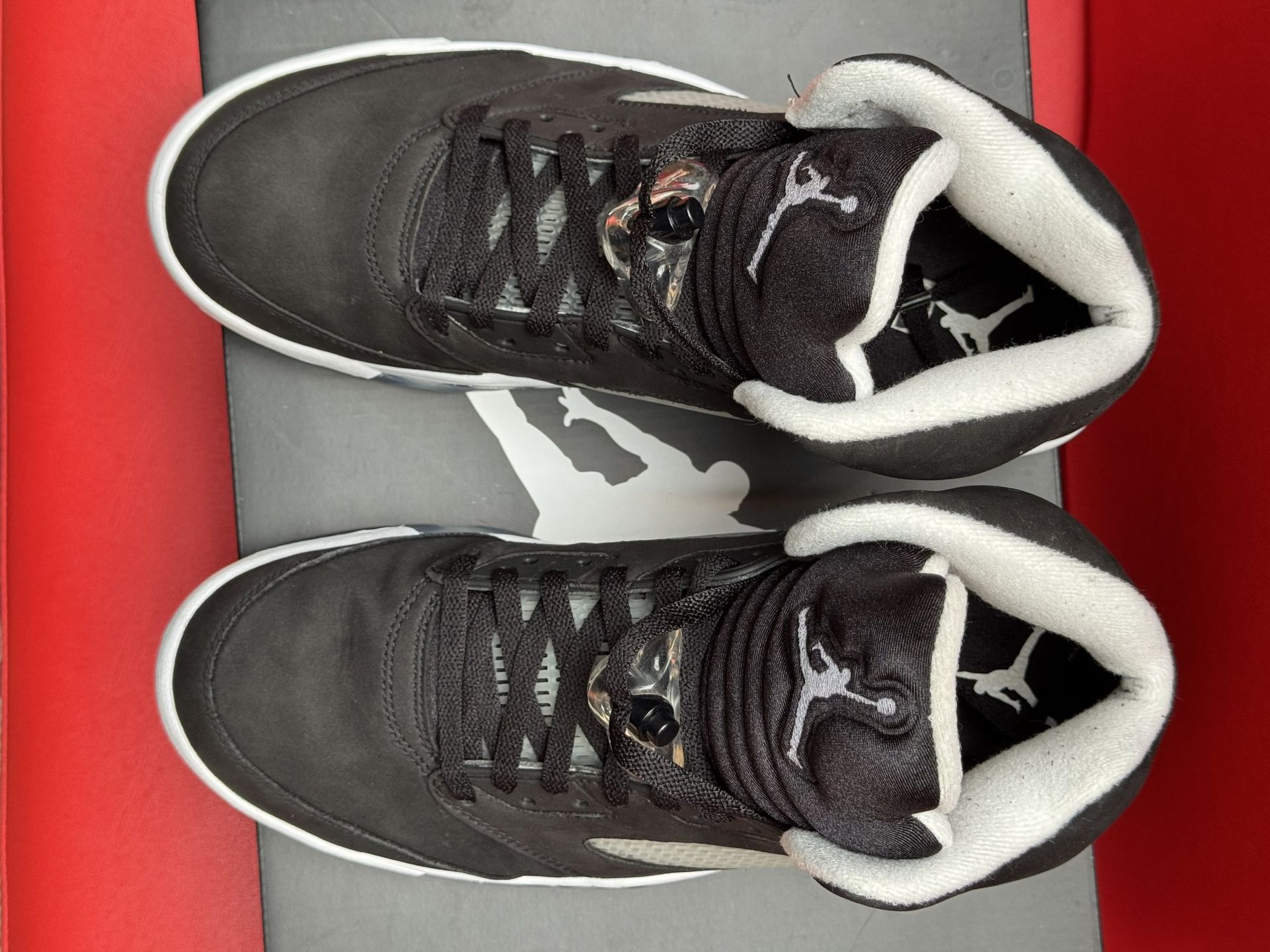 Black and white Air Jordan 5 sneakers with a clear sole, sitting on a black shoebox, against a red background.