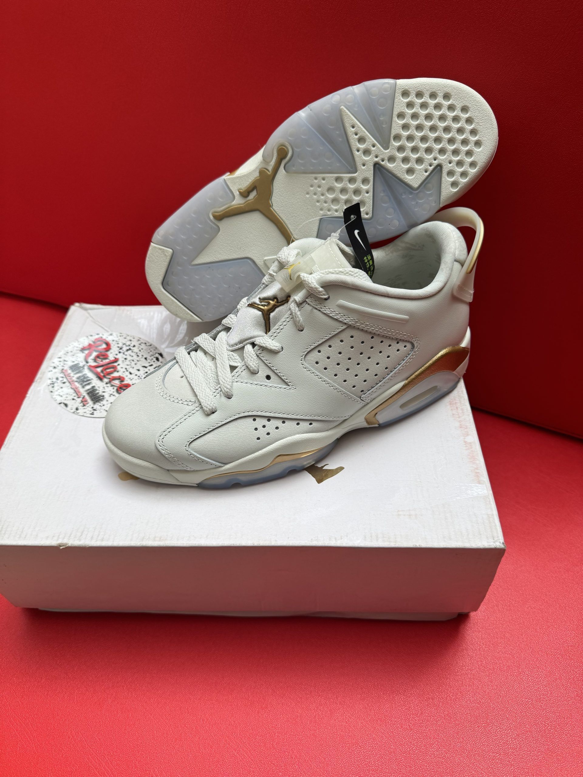 White and gold Air Jordan 6 sneakers on a white box, one turned over showing the sole, against a red background.