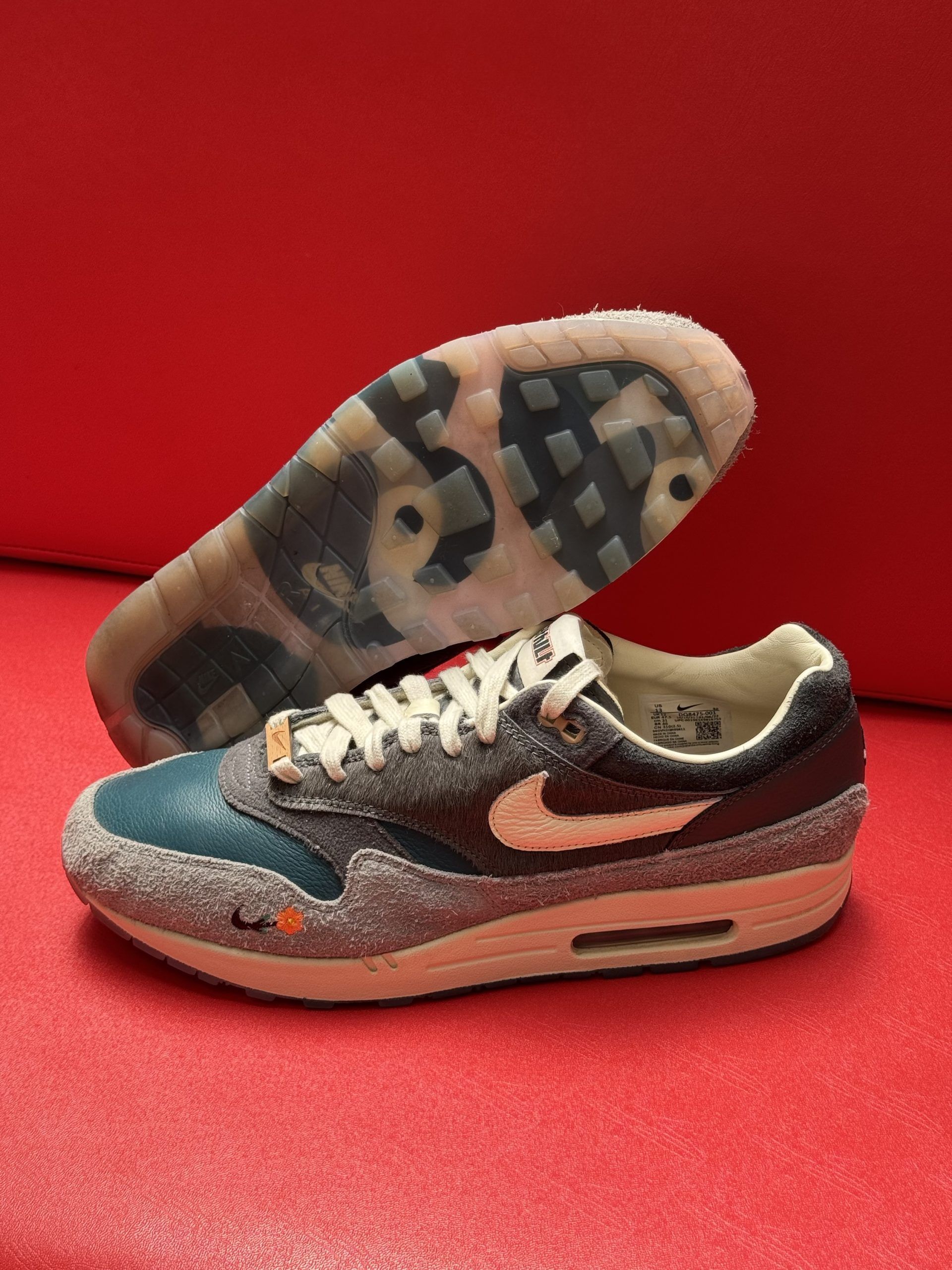 Nike Air Max 1 sneaker with a blue, gray, and cream color scheme, with a clear sole on a red surface.