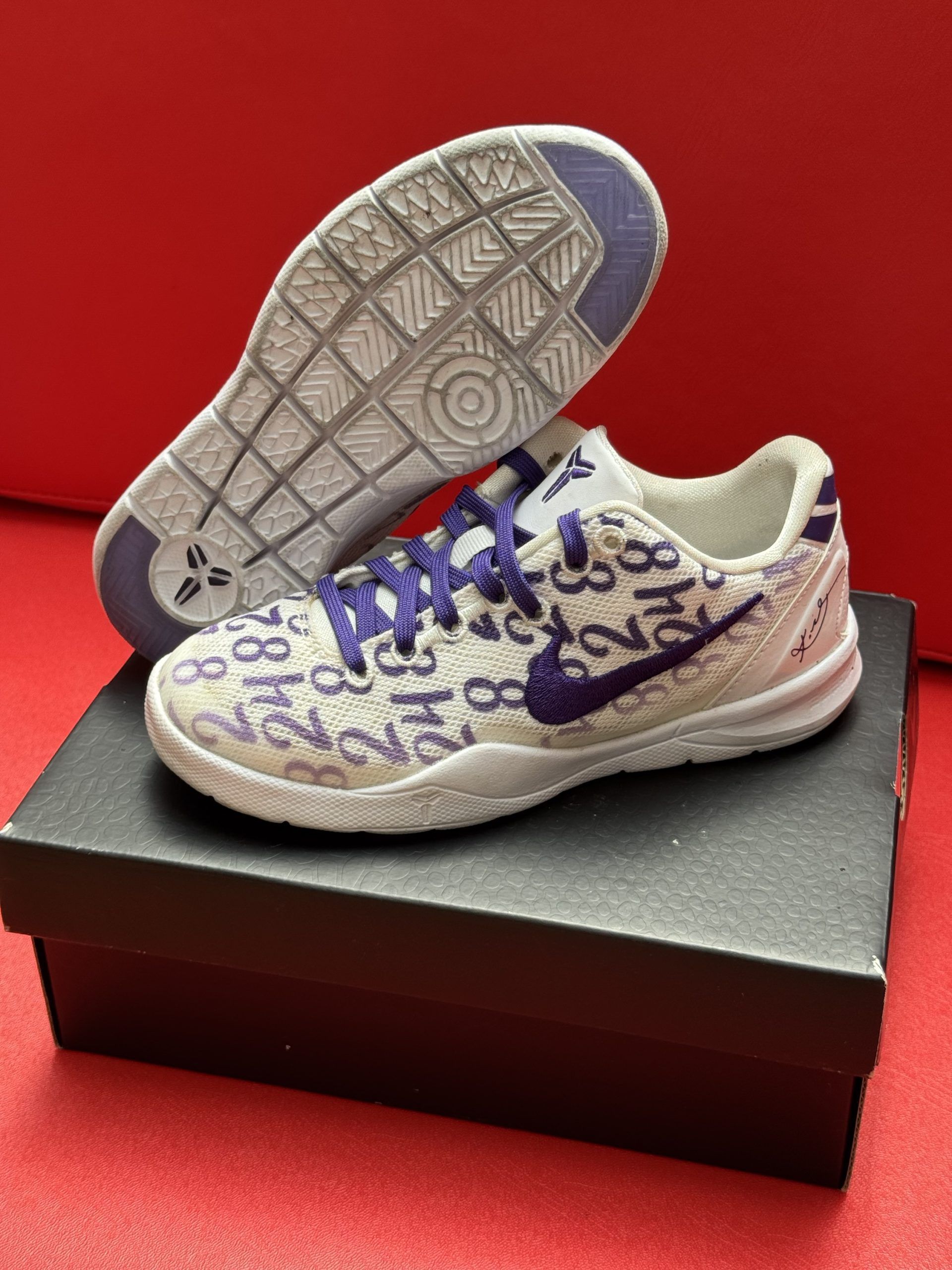 Nike Kobe 6 sneakers, white with purple accents, displayed on a black box, against a red background.