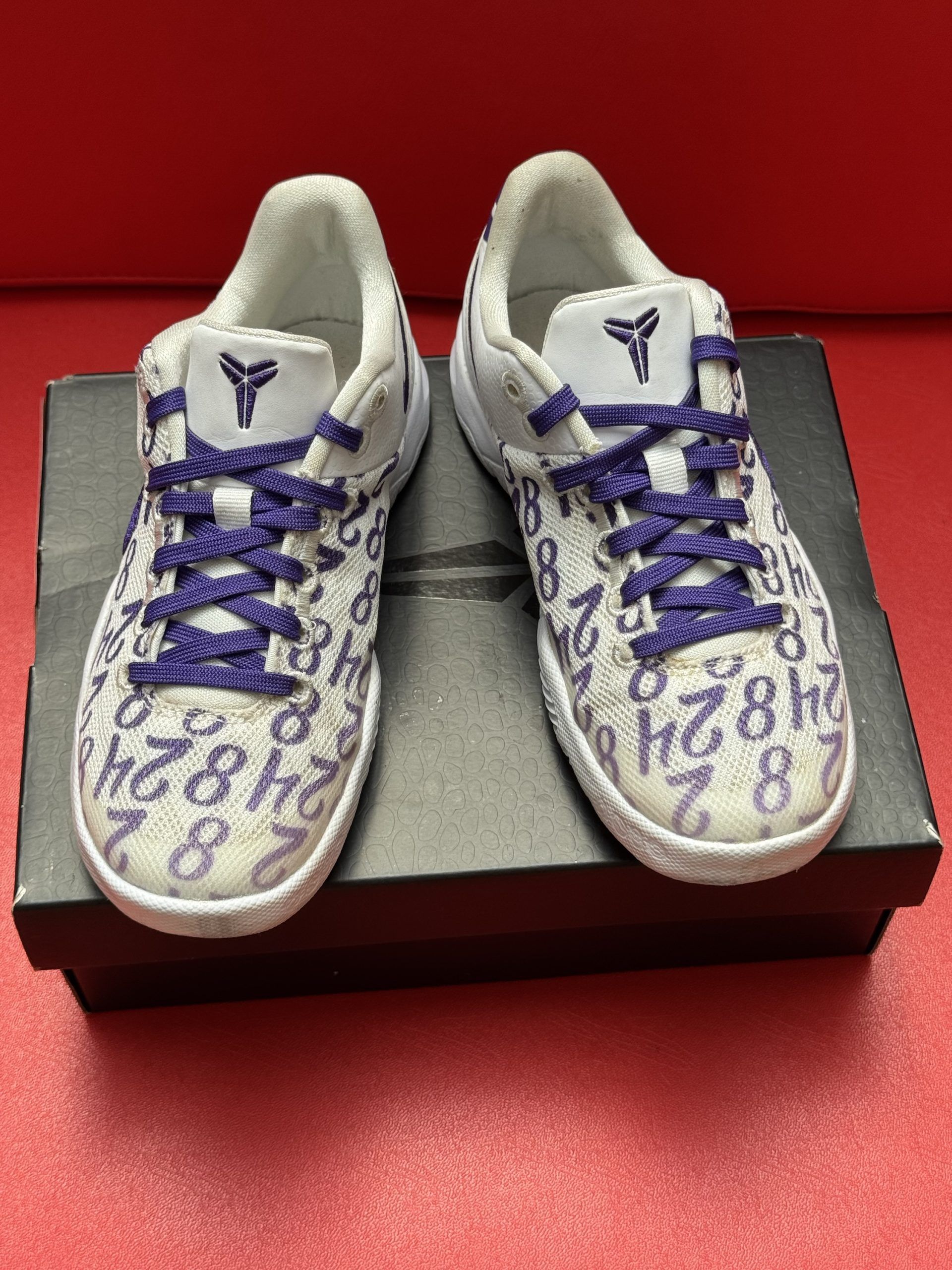 Nike Kobe 6 sneakers, white with purple accents, displayed on a black box, against a red background.