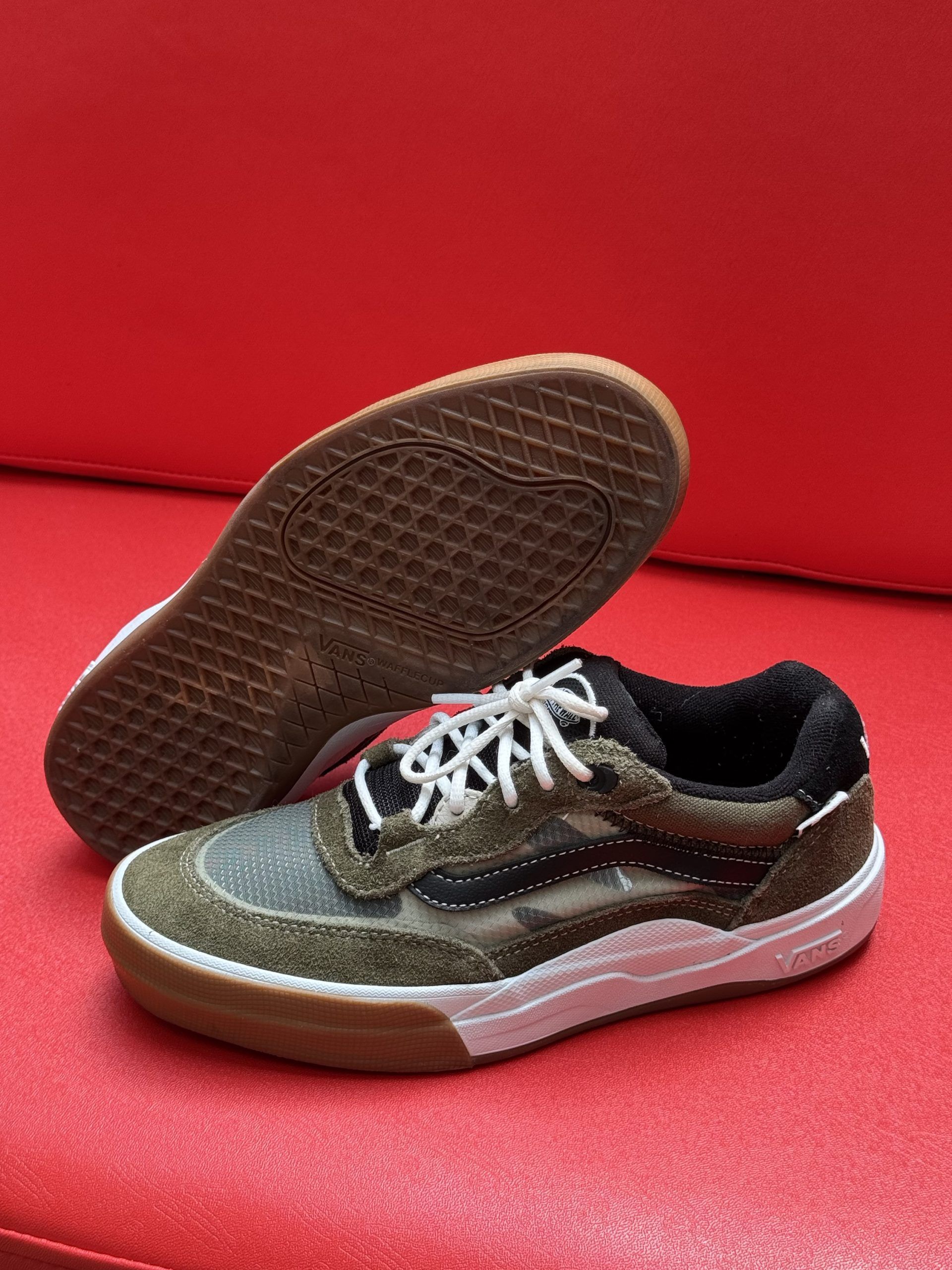 Tan and black Vans sneakers with white laces, brown sole, and white trim on a red surface.