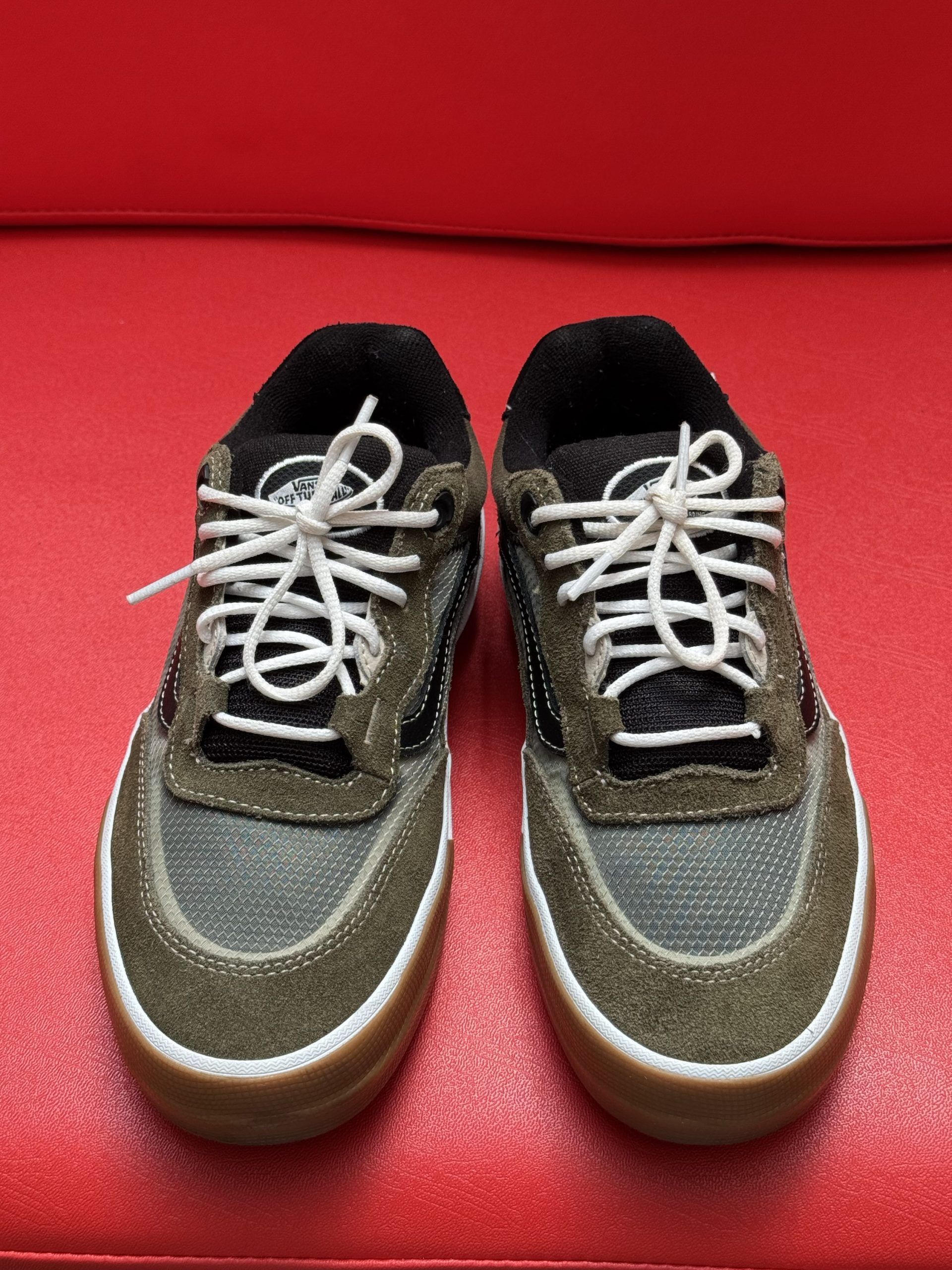 Tan and black Vans sneakers with white laces, brown sole, and white trim on a red surface.