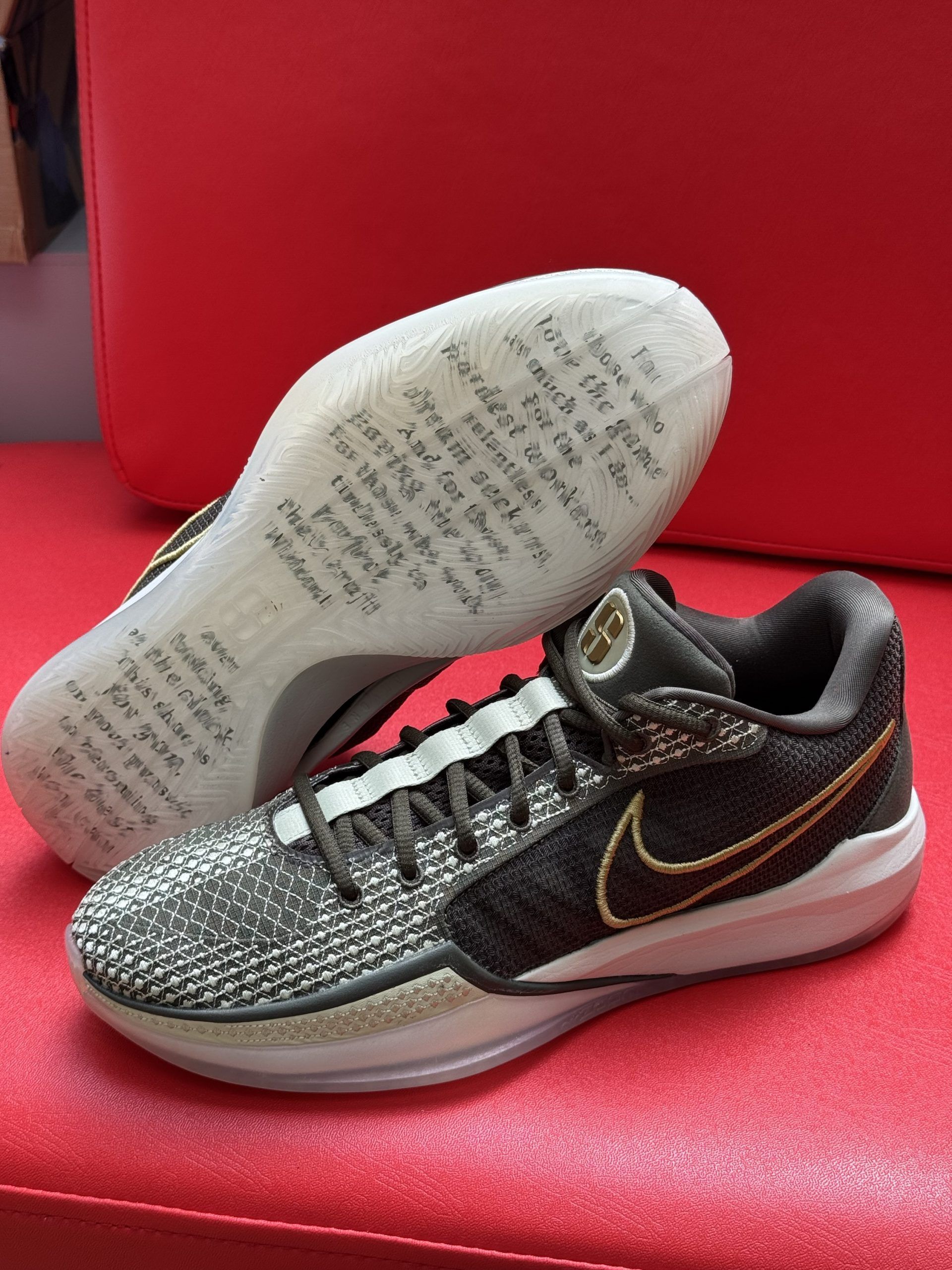 Pair of black and gold Nike Kobe basketball sneakers on a red surface, with text on the translucent soles.