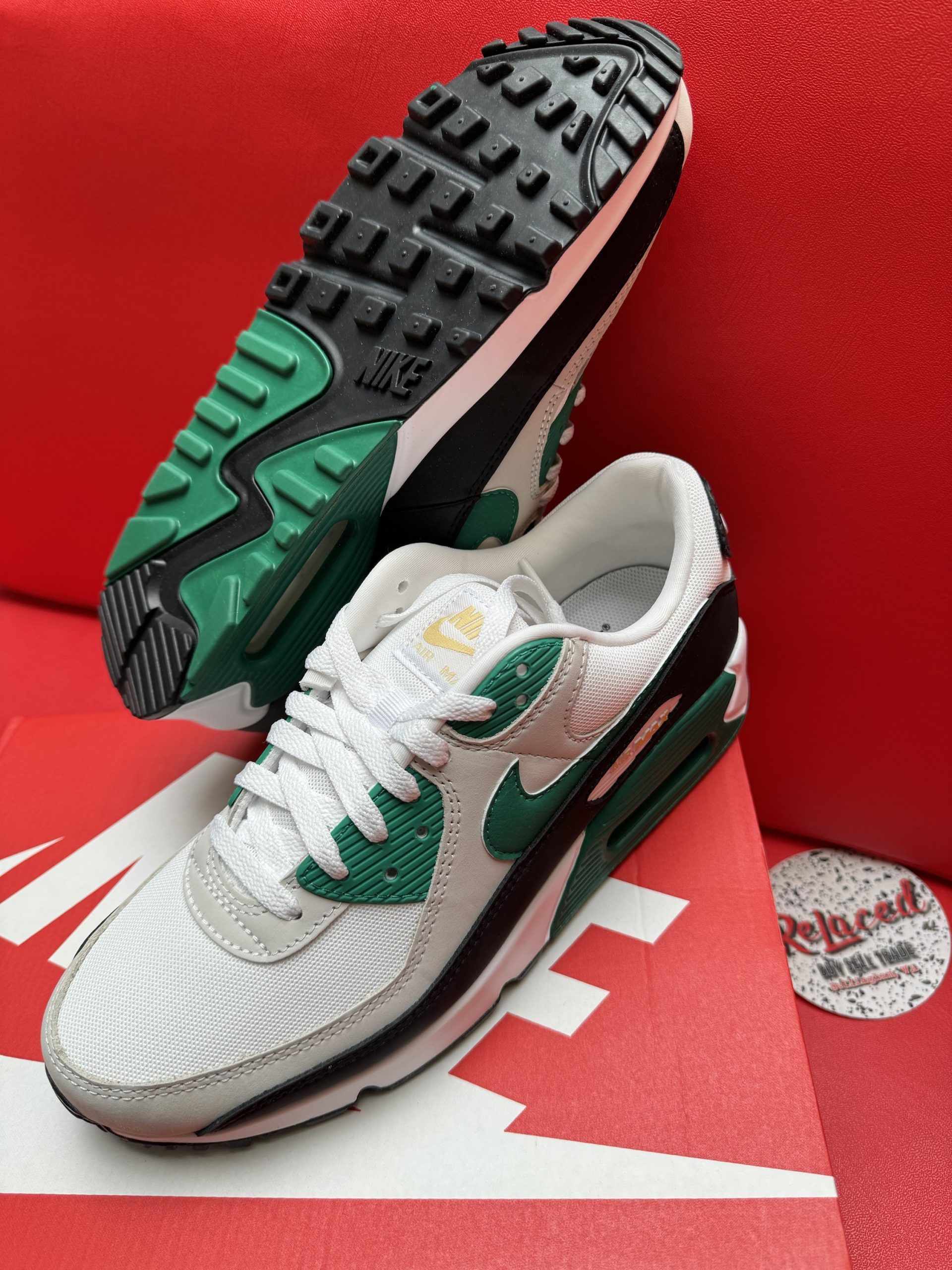 Pair of Nike Air Max 90 sneakers, white, green, and gray, on a red shoebox, sole up.