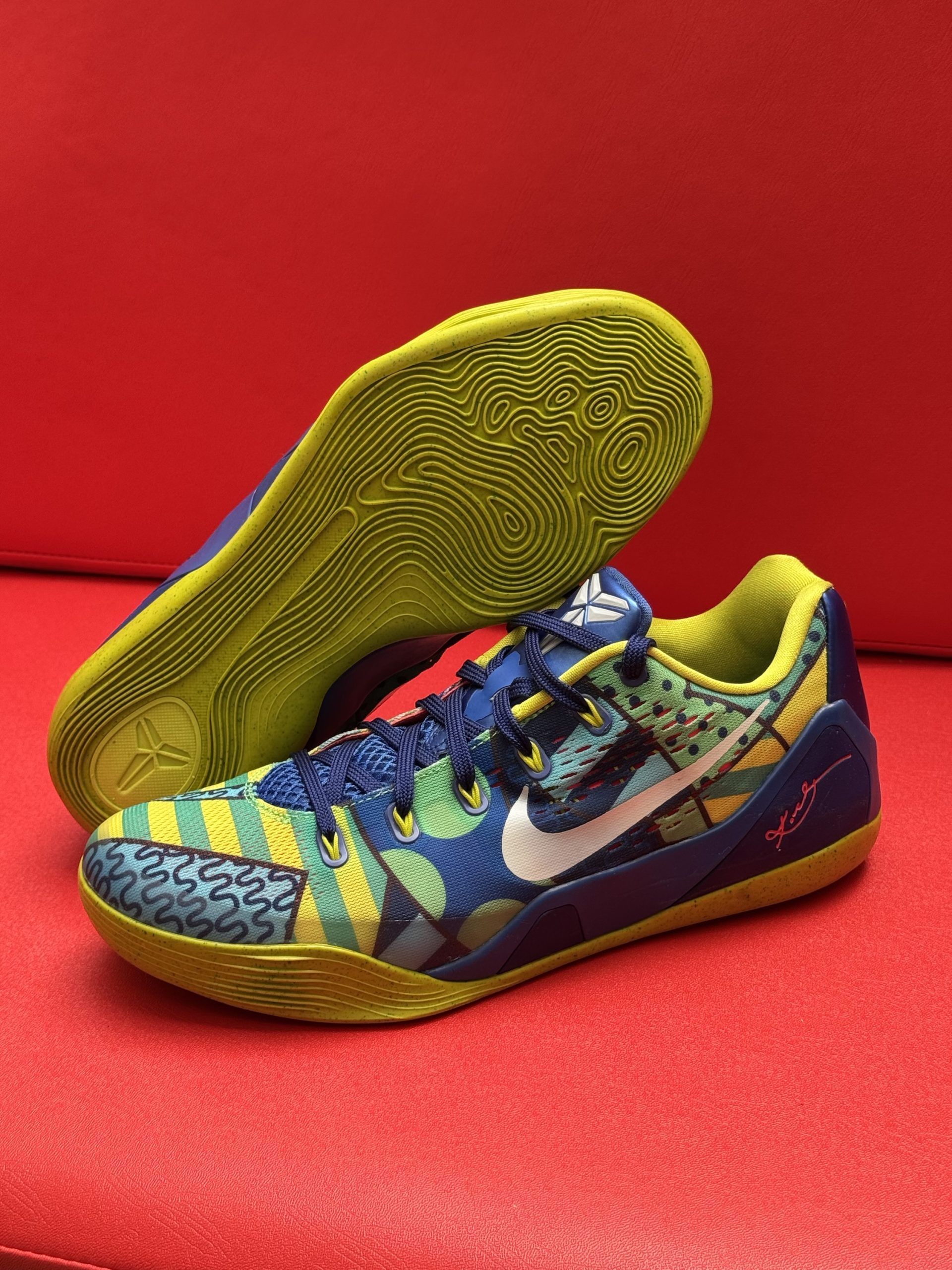 Blue, yellow, and green Kobe basketball shoes on a red surface.