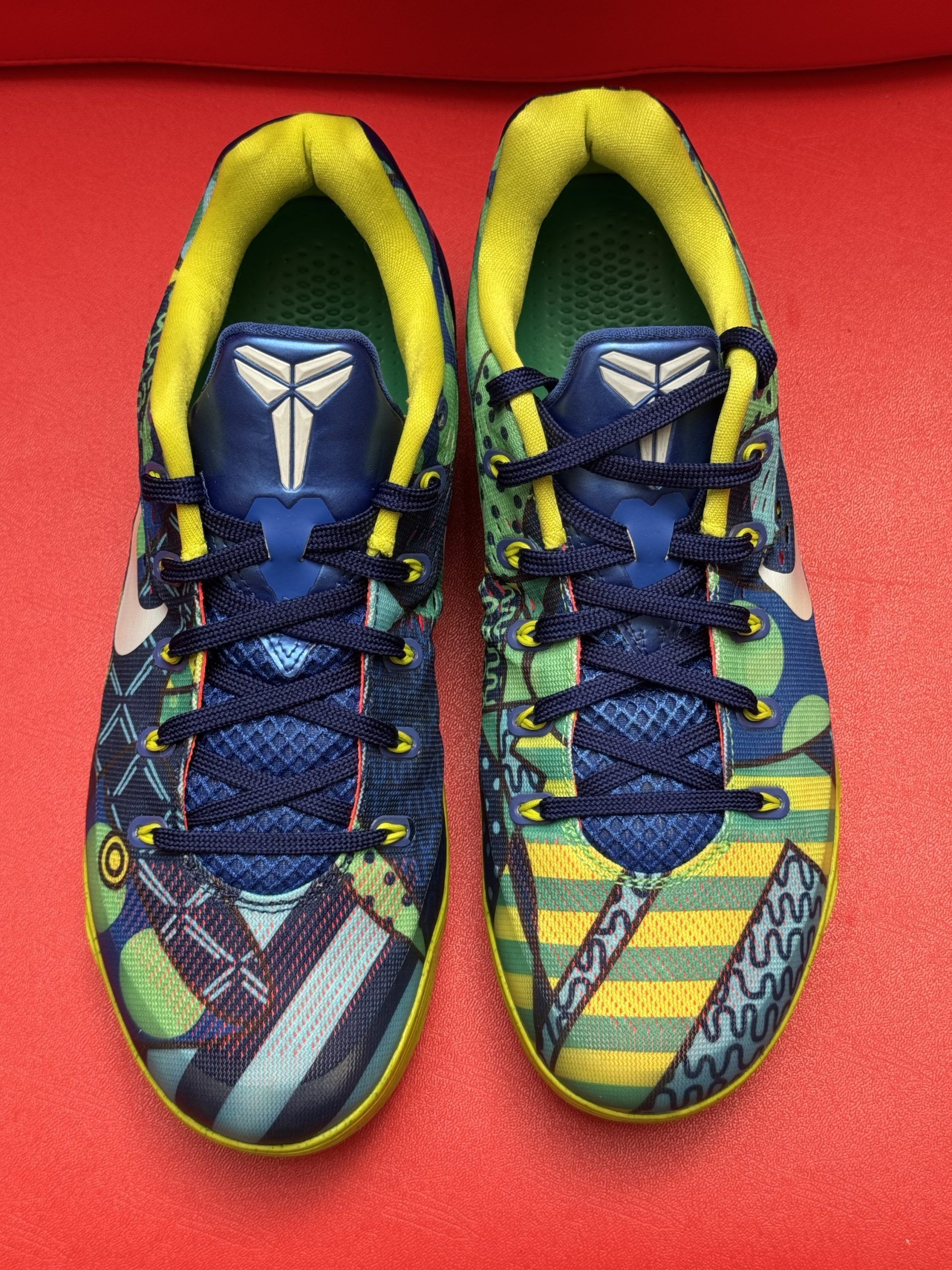 Blue, yellow, and green Kobe basketball shoes on a red surface.