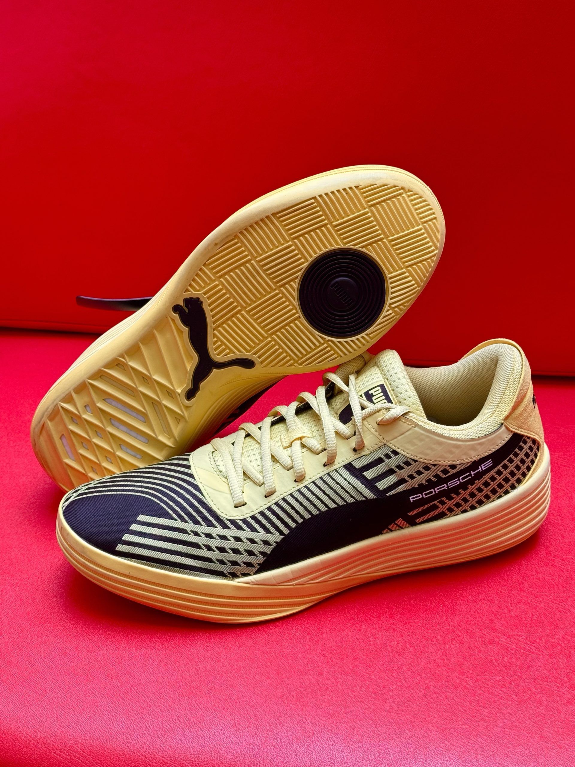 Puma sneakers with beige sole, yellow laces, and black and blue patterned upper on red surface.