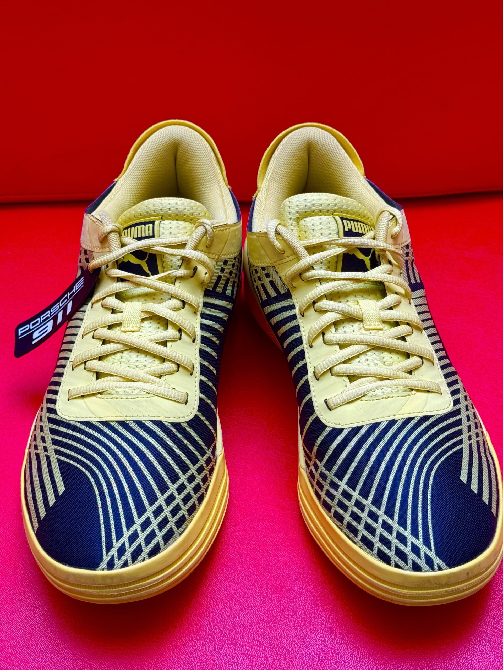 Puma sneakers with beige sole, yellow laces, and black and blue patterned upper on red surface.