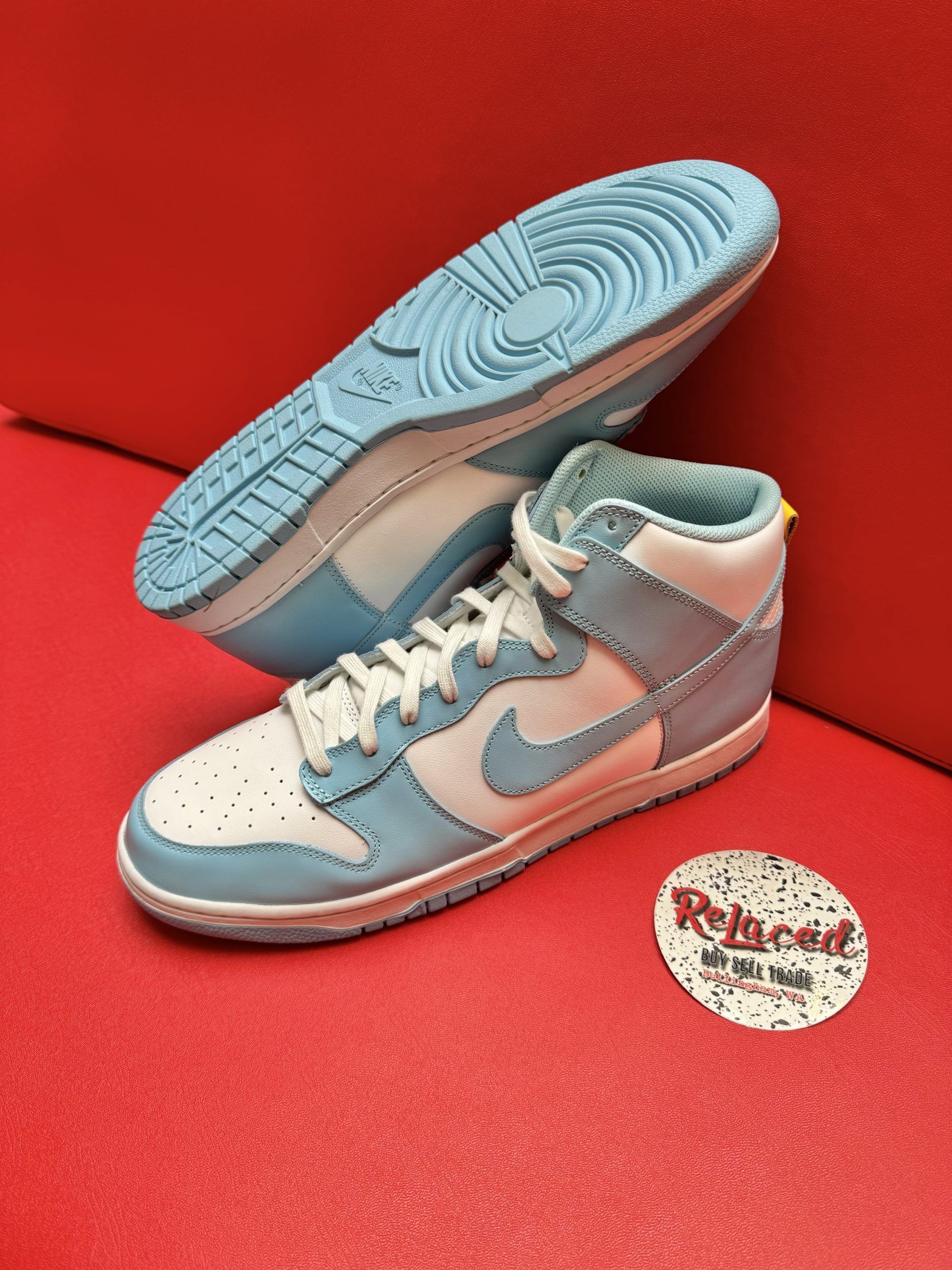 Pair of light blue and white Nike Dunk High sneakers on a red surface, with a circular sticker.