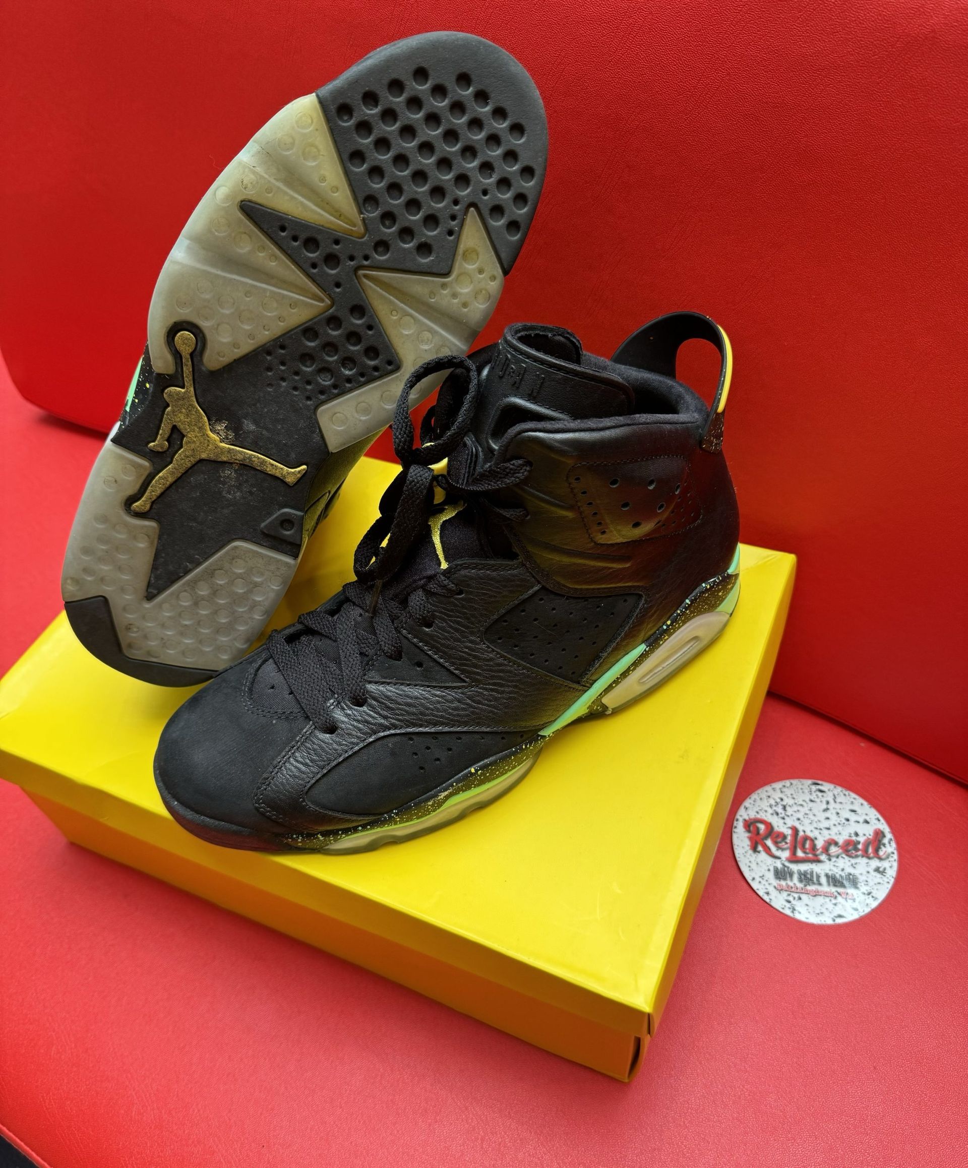 Black and yellow Air Jordan 6 sneakers on a yellow box, against a red backdrop. The sole shows a gold Jumpman logo.