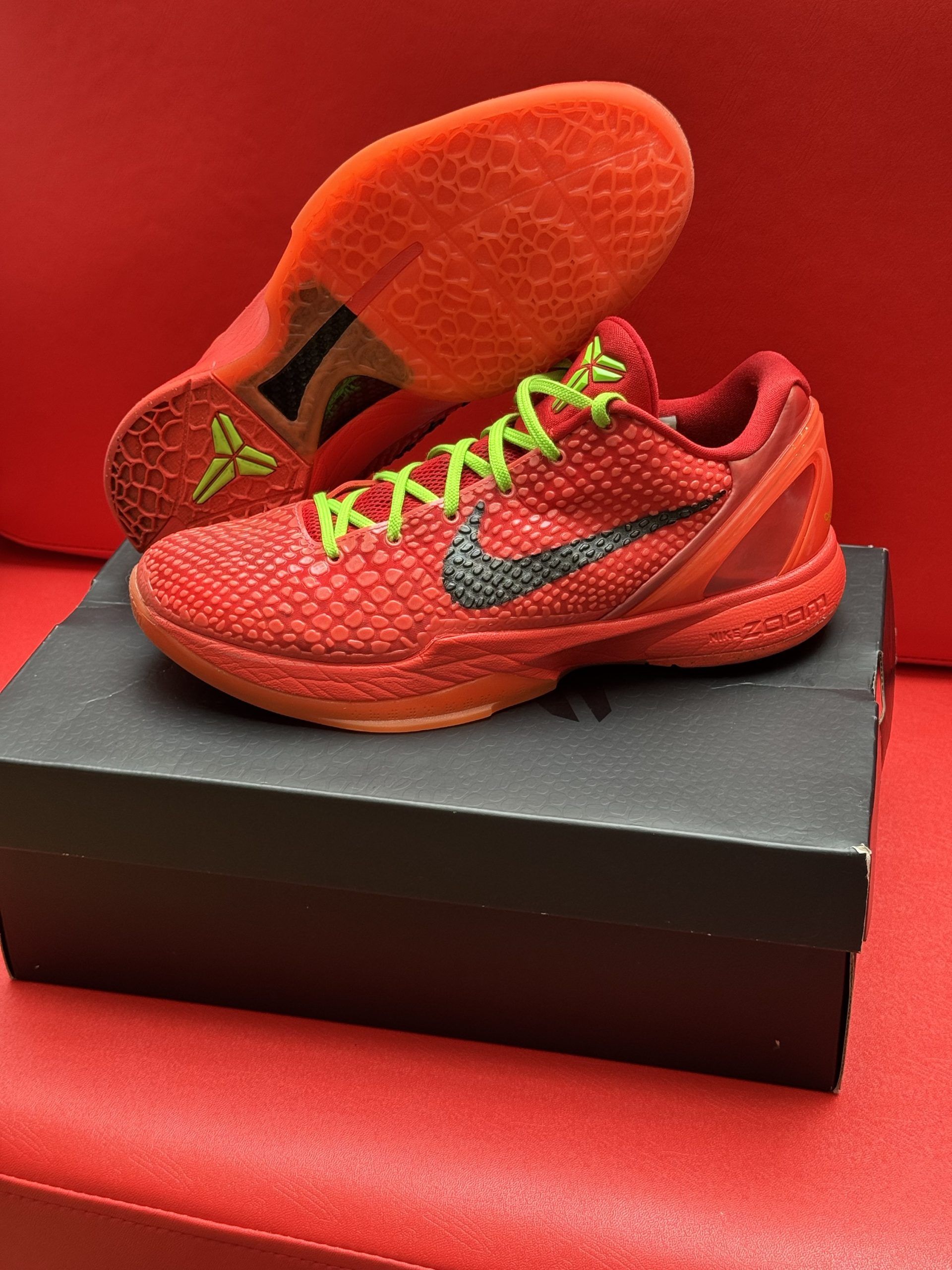 Red Nike Kobe basketball shoes with black swoosh and green laces sit on a box, red background.