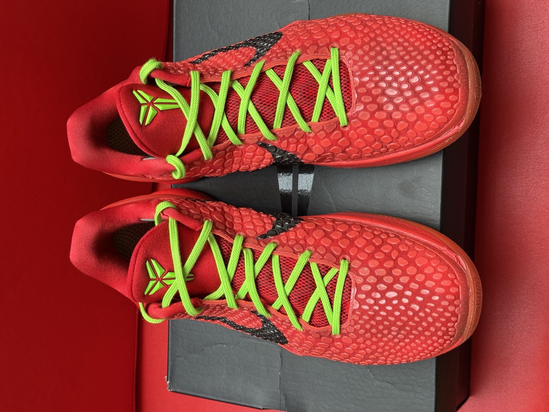 Red Nike Kobe basketball shoes with black swoosh and green laces sit on a box, red background.