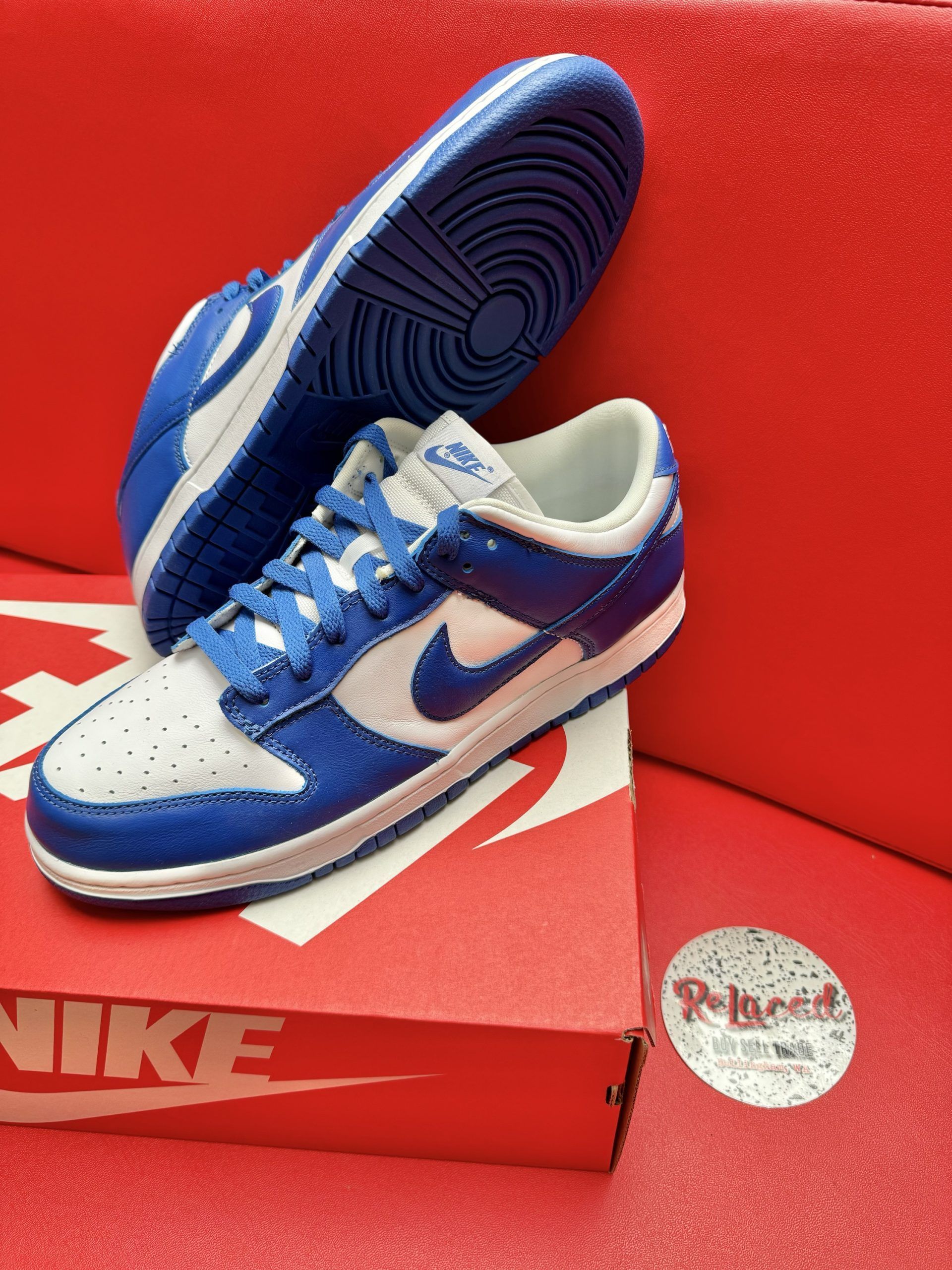 Blue and white Nike Dunk Low sneakers, one on its side, atop a red shoebox on a red surface.