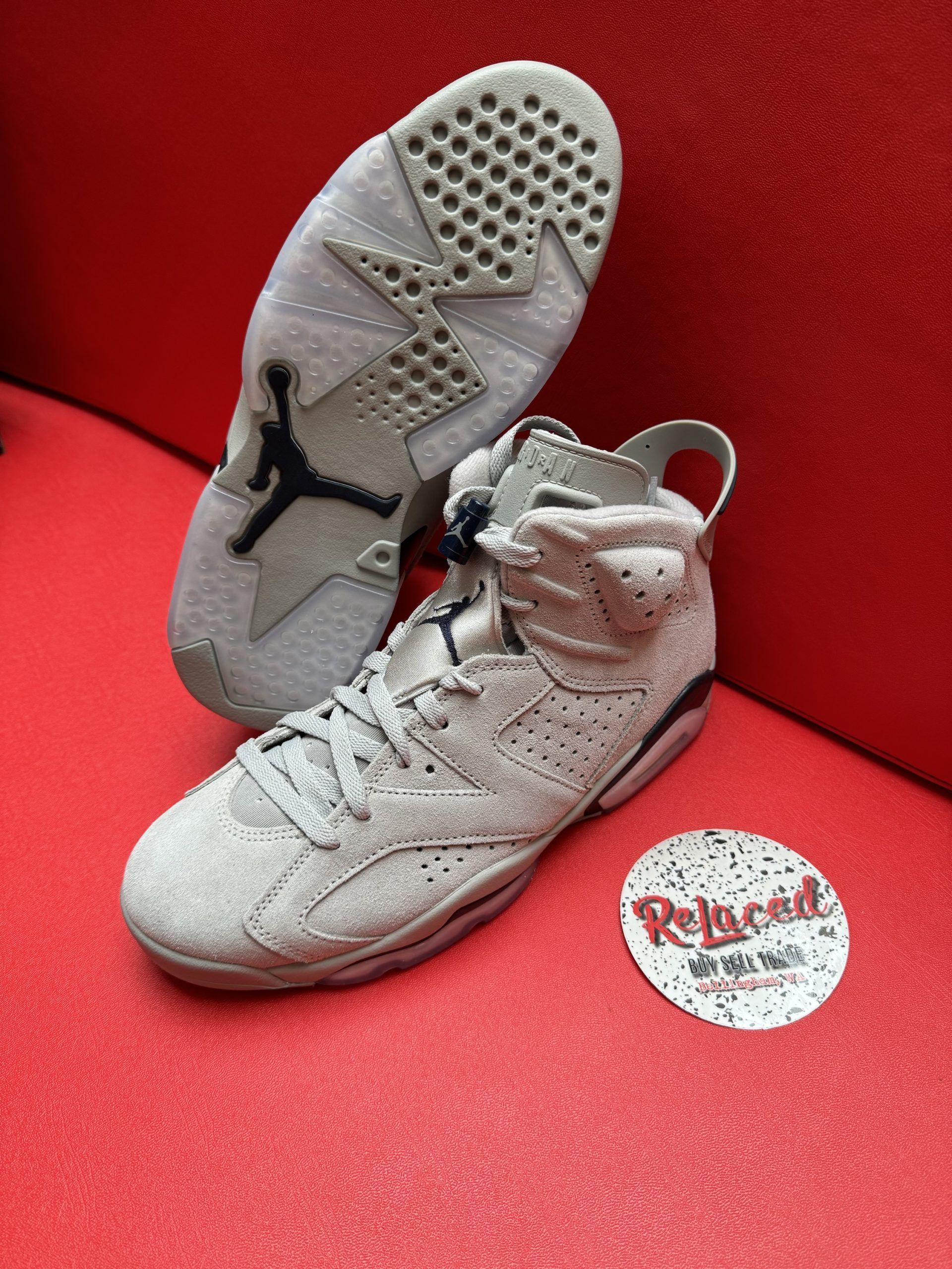 Air Jordan 6 sneakers, light grey suede, one sole up, on a red surface with a logo.