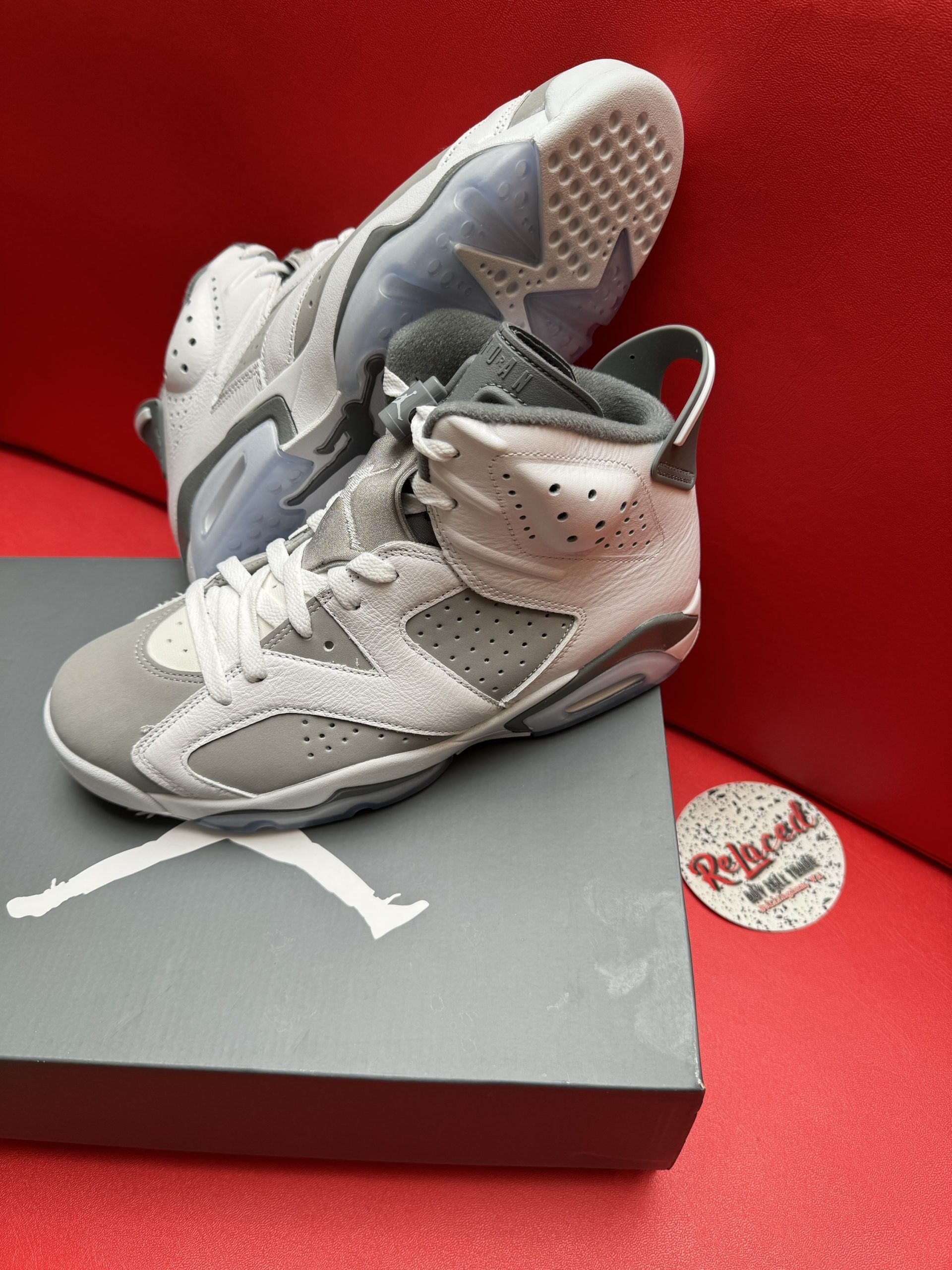 Air Jordan 6 sneakers, gray and white, on top of a gray Jordan shoe box against a red background.