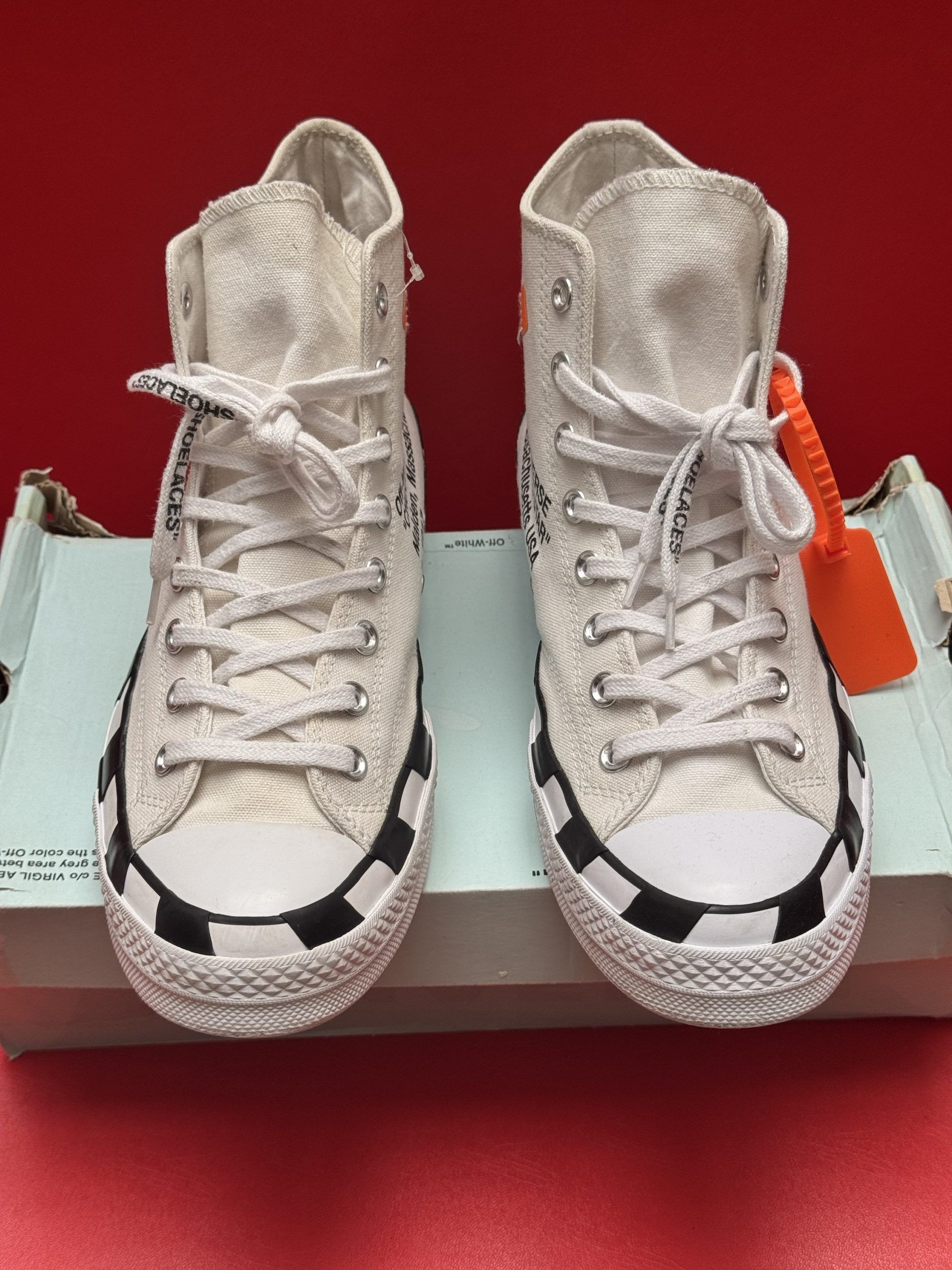 Off-White Converse sneakers, white canvas, black striped soles, black text, with box, against red background.