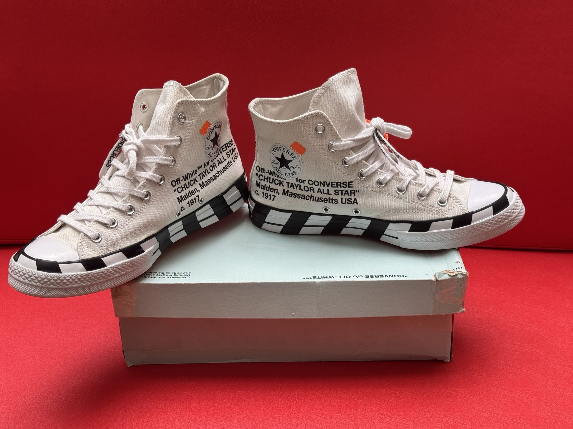 Off-White Converse sneakers, white canvas, black striped soles, black text, with box, against red background.