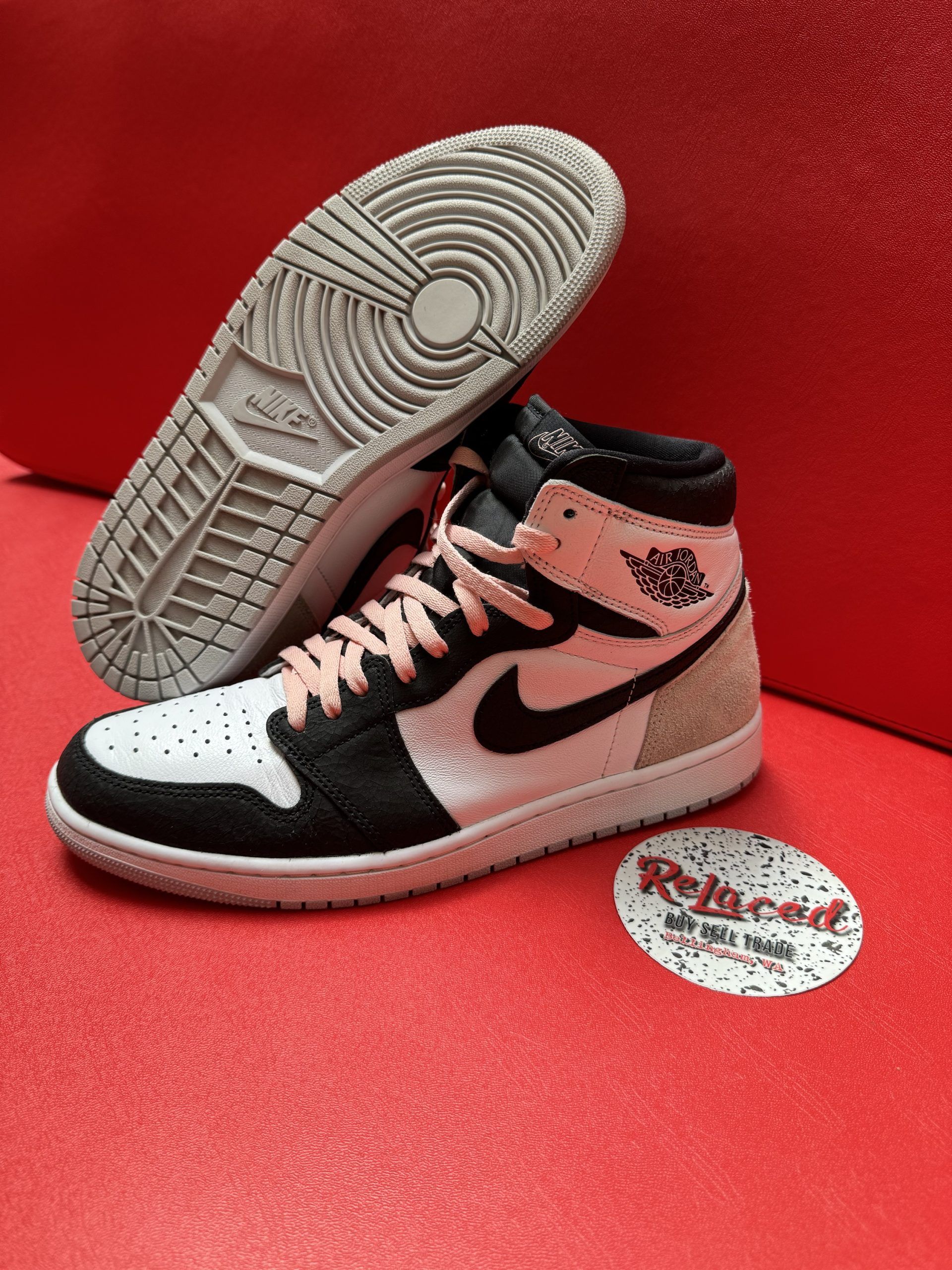 Air Jordan 1 High sneaker, white and black with pink laces and beige accents, set on a red surface.