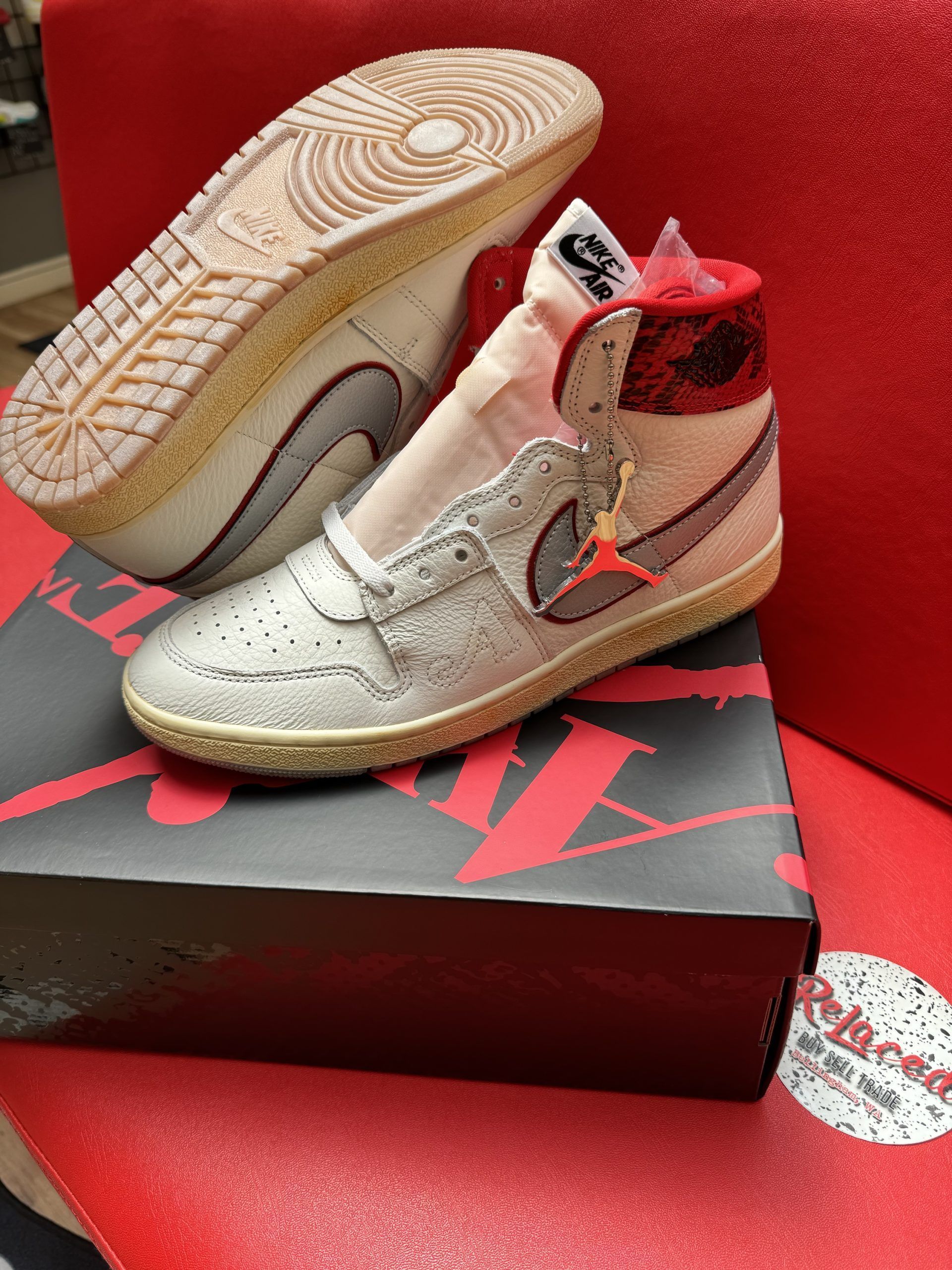 Air Jordan 1 High sneakers on a black shoe box, set on a red surface. One shoe is upright, the other’s sole facing upward.