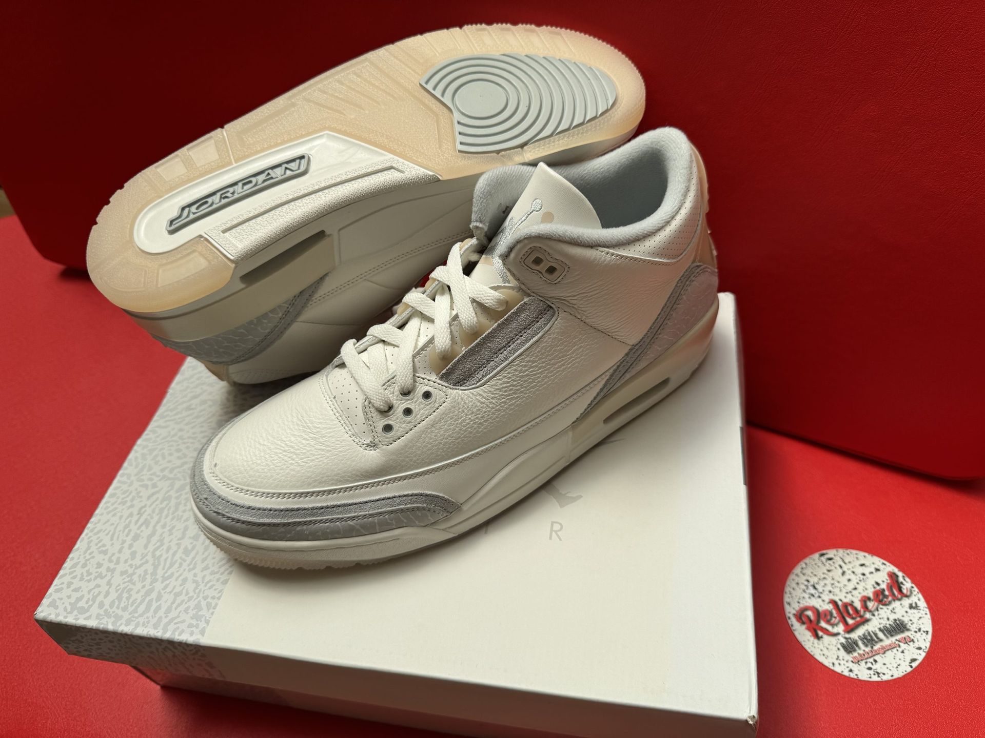 Off-white and gray Air Jordan 3 sneakers on a white box, with a red background. One shoe is flipped to show the sole.