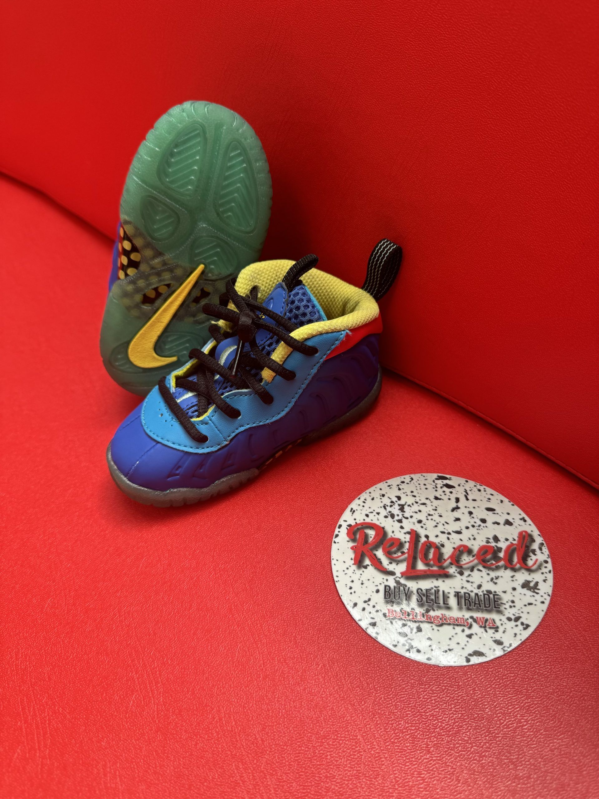 Miniature blue, yellow, and red sneaker with green sole, next to a Relucaed logo, on a red surface.