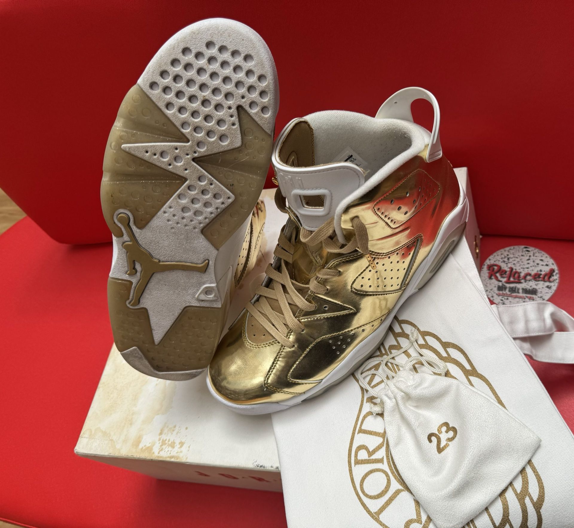 Gold and white Air Jordan 6 sneakers, displayed on a box with a white bag and red surface.