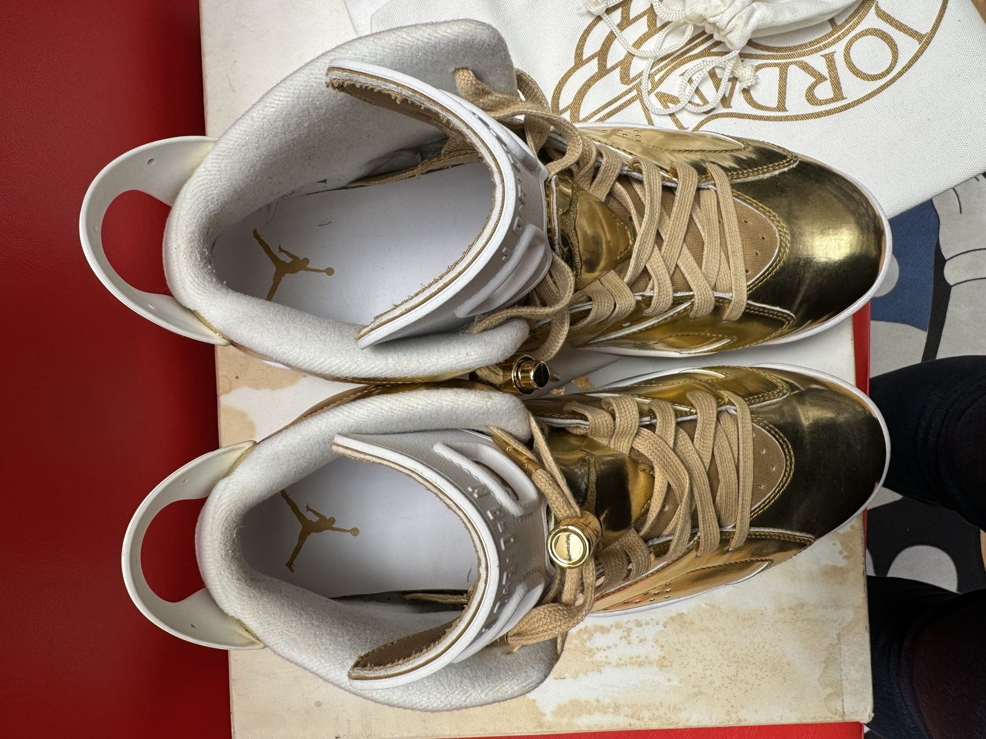 Gold and white Air Jordan 6 sneakers, displayed on a box with a white bag and red surface.