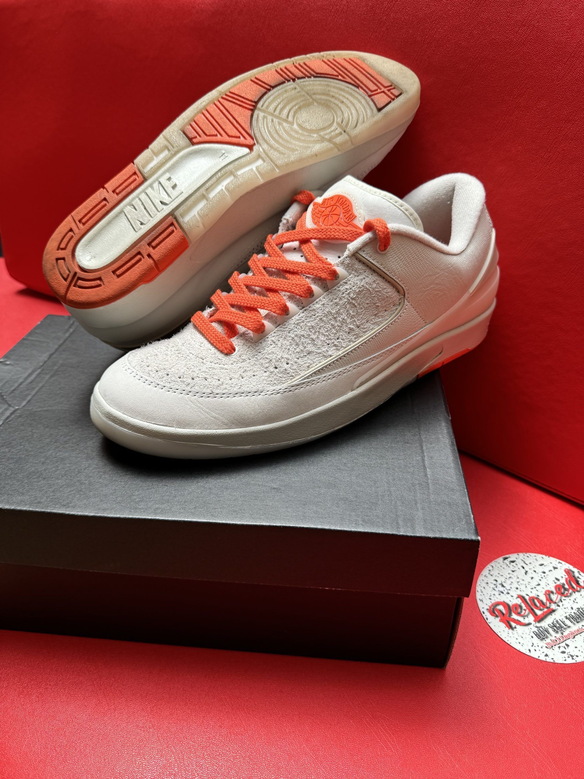 Pair of light gray sneakers with orange laces, one shoe upside down showing the sole. Shoes sit atop a black box on a red surface.